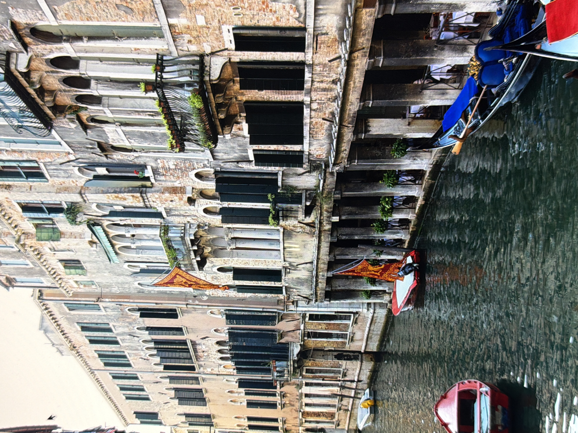 The image depicts a canal in Venice, Italy. The canal is bordered by tall, old buildings with Venetian architecture. The buildings feature arched windows, balconies with potted plants, and weathered facades. Several boats are visible in the canal. In the foreground, a red boat is docked. Further down the canal, a red and white boat with an outboard motor is also present. To the right, a black gondola with blue seating is tied up. The water in the canal is dark and reflective. The overall impression is one of a traditional Venetian waterway scene.