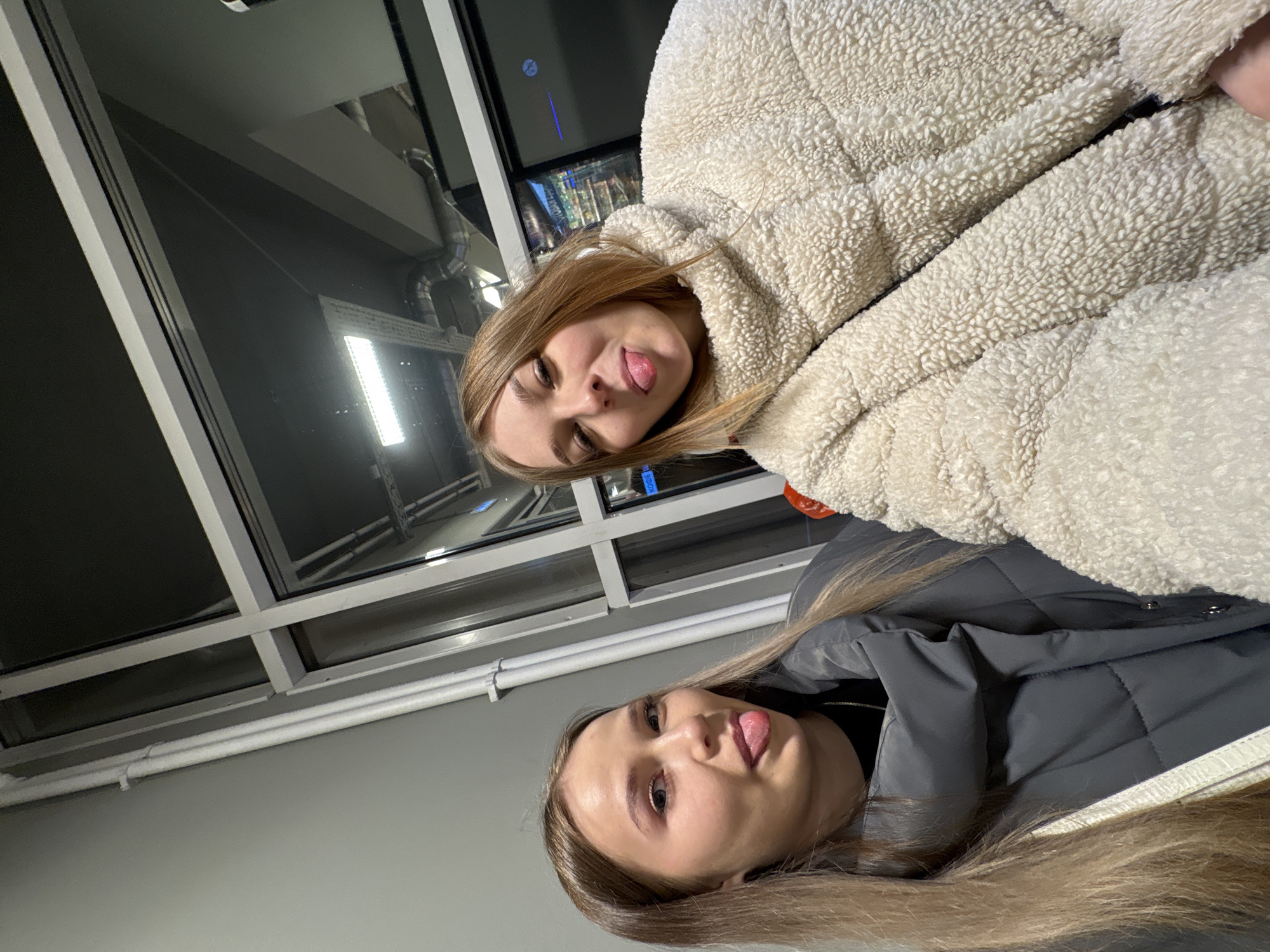 Two young women are captured in a playful, rotated portrait, seemingly taken from a low angle in an indoor setting. Both subjects have long, light brown hair and are sticking out their tongues, indicating a lighthearted and casual mood.
The woman positioned higher in the frame is wearing a cream-colored, fluffy, possibly sherpa-style jacket. Her companion, lower in the frame, is dressed in a dark gray quilted puffer jacket.
The background features modern architectural elements, including large glass panels or windows. Through or reflected in these panels, industrial-style components like exposed pipes or ventilation ducts and bright, rectangular light fixtures are visible. A white pipe runs horizontally along the lower part of the window structure. The overall lighting suggests an interior environment, with the darkness in the reflections possibly indicating evening or night outside.
A small, blue label with the text "KODE" is visible in the background, appearing vertically oriented near the lower woman's shoulder and faintly near the upper woman. The scene takes place in what appears to be a public or commercial building in Chaikovsky, Russia.