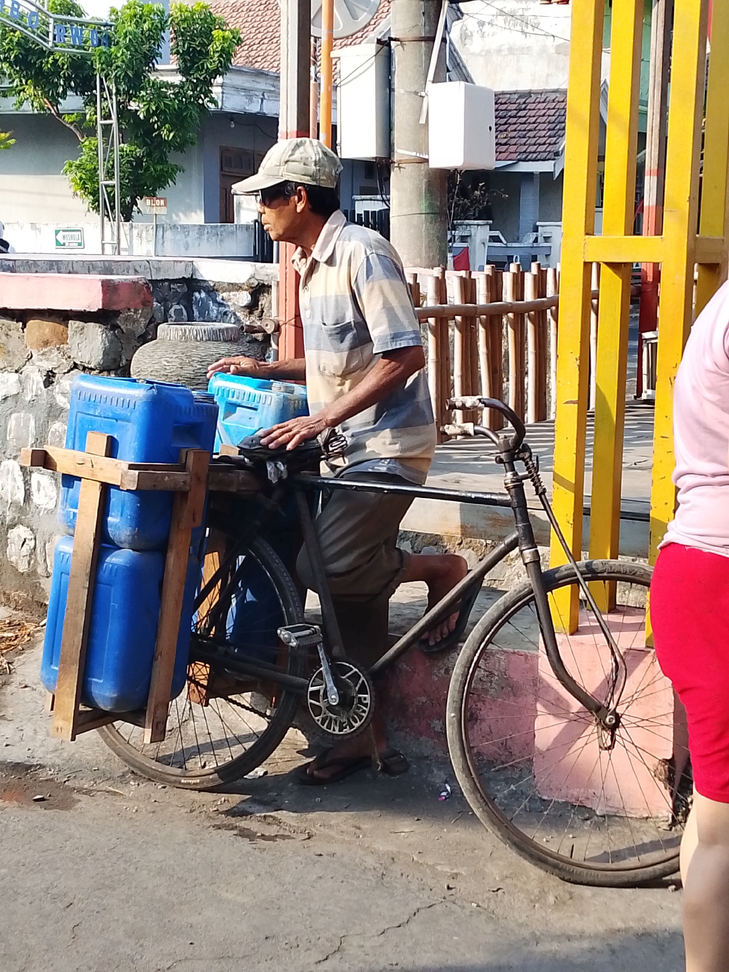 The image depicts a street scene in the city of Kediri, Indonesia. In the foreground, a man wearing a cap, sunglasses, and a striped short-sleeved shirt is sitting on a bicycle. The bicycle is loaded with several large blue plastic containers, secured with wooden supports. The man's hands are on the handlebars. To the right of the bicycle, part of another person wearing a pink shirt and red pants is visible. The background shows buildings with tiled roofs, a bamboo fence, and a bright yellow structure. The scene appears to be outdoors, with sunlight casting shadows.