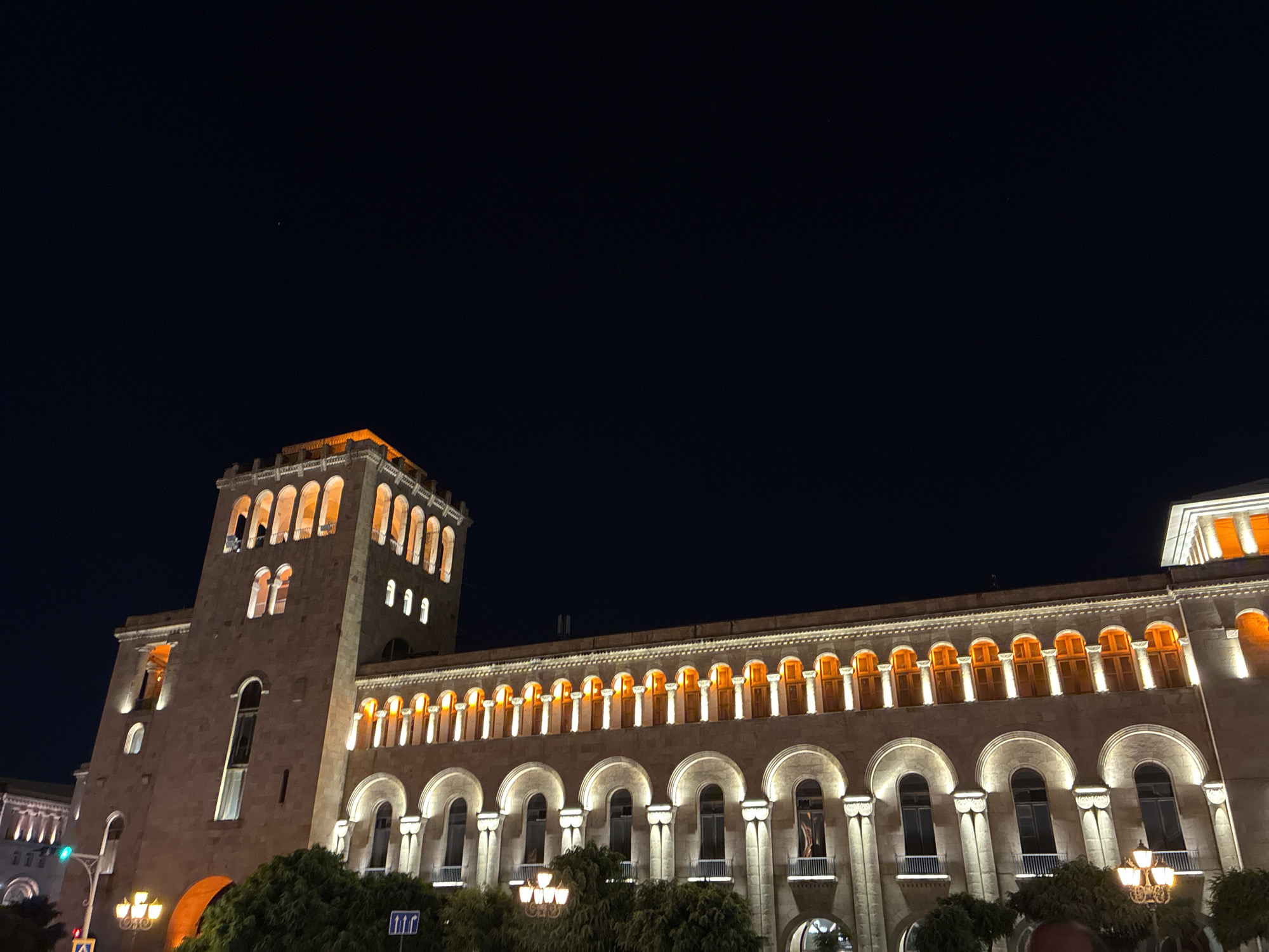 A wide, low-angle shot captures a large, stone building illuminated at night against a dark sky. The building features a prominent tower on the left, with arched windows lit by a warm orange glow. A long facade stretches across the center and right of the frame, characterized by a repeating pattern of arched windows and columns, also lit with orange light. Trees are visible at the base of the building, and streetlights and traffic signals can be seen on the lower left. The sky is almost completely black, with a faint hint of a distant star visible in the upper left quadrant. The overall impression is one of historical architecture beautifully lit for the evening.