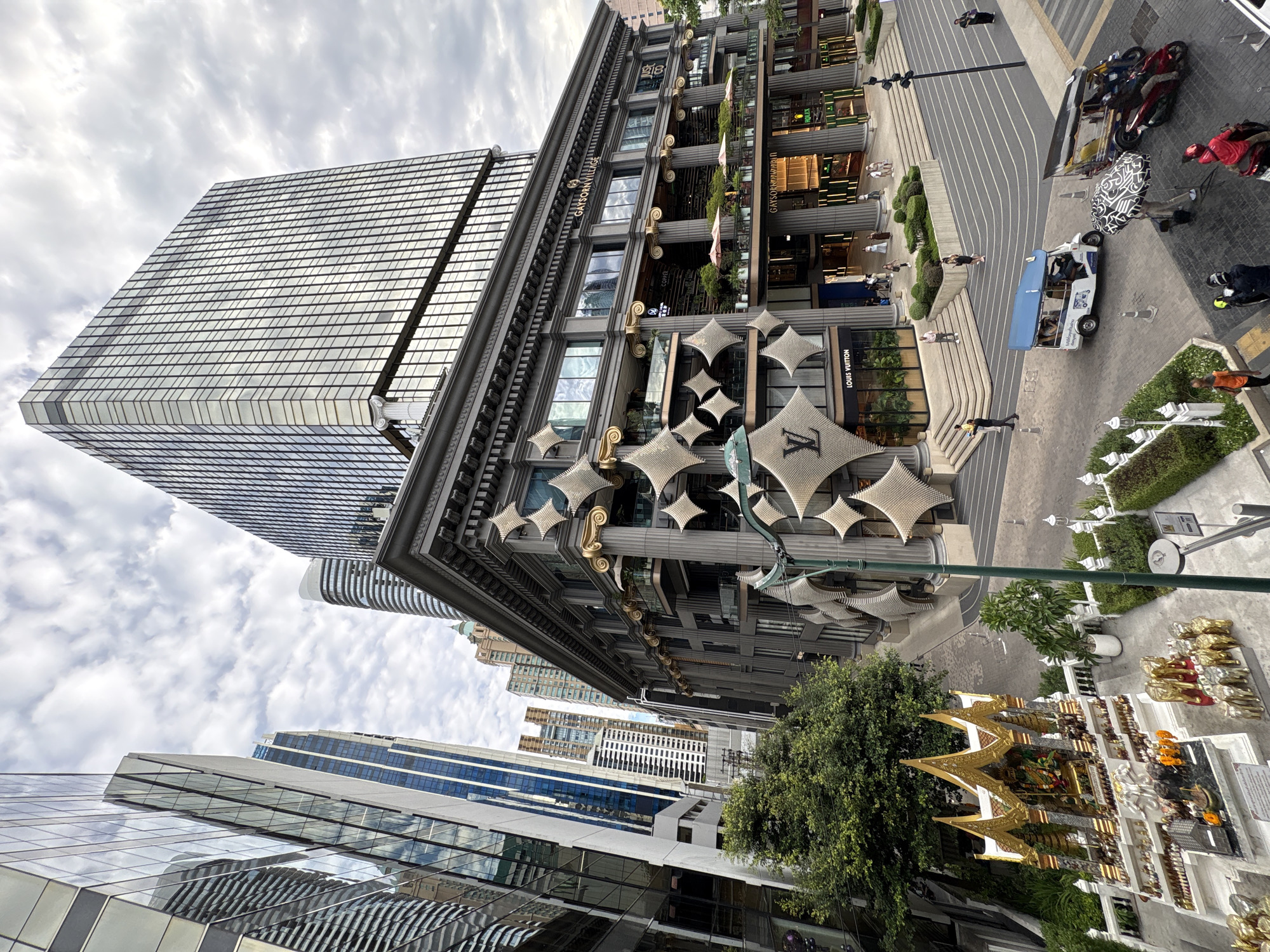 The image depicts a street scene in Bangkok, Thailand, with a variety of architectural styles on display. At the forefront is a traditional Thai spirit house, decorated with gold accents and figurines. A large tree partially obscures the spirit house. Beyond the tree, a modern building adorned with diamond-shaped ornaments and Louis Vuitton logos stands prominently. Adjacent to this building is a taller, grid-patterned skyscraper.
In the background, a mix of high-rise buildings of varying designs rises into a cloudy sky. The street-level scene includes pedestrians walking on a paved area with curved stripes, a tuk-tuk parked on the road, and several motorcycles. The scene is captured from a low angle, looking upward at the buildings.