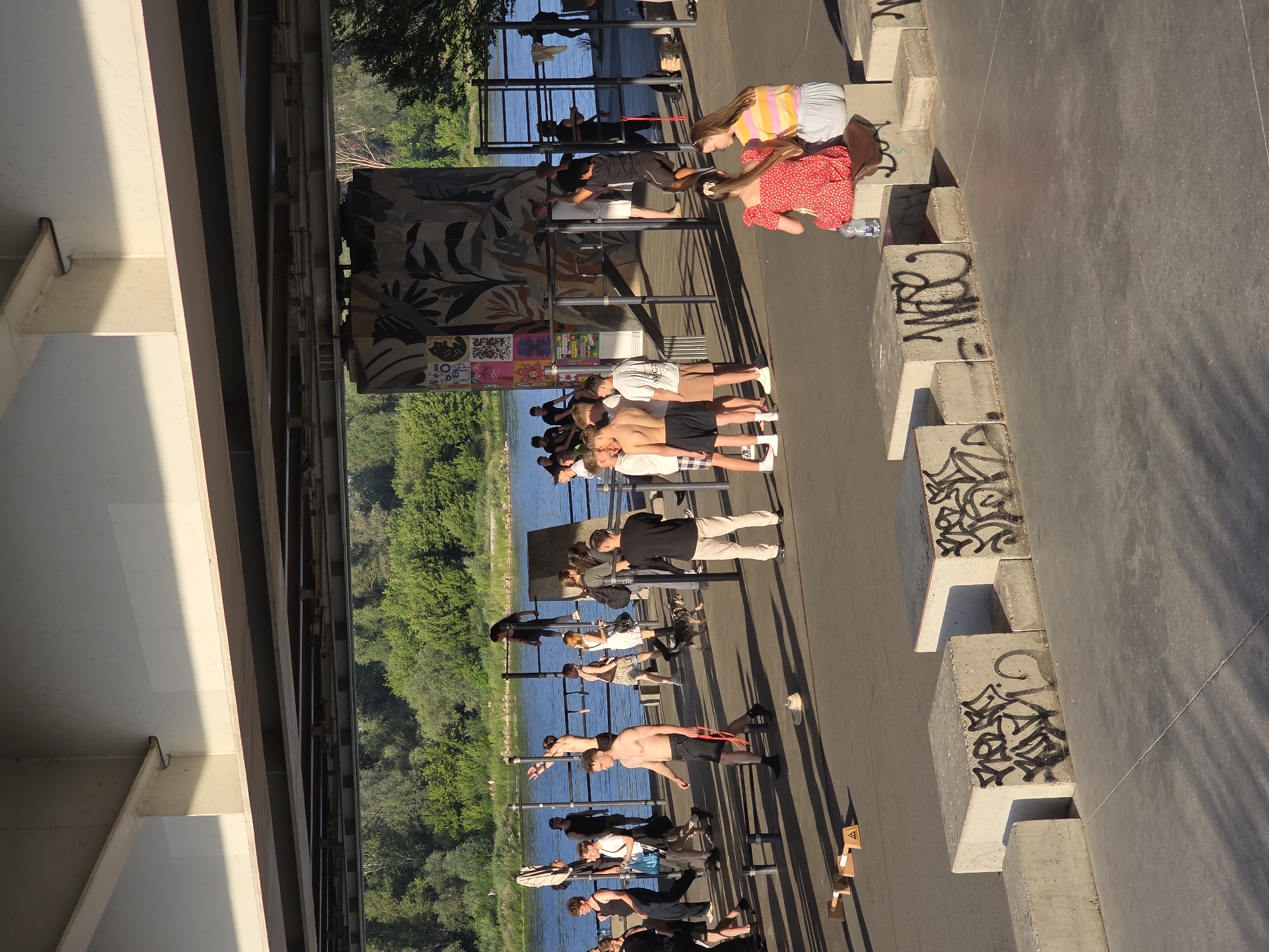 The image captures a sunny day at a Warsaw, Poland outdoor fitness area situated beneath a bridge. The foreground is dominated by concrete structures, some bearing graffiti, and shadows cast by the bridge overhead. A group of young people are engaged in various activities: some are working out on pull-up bars and parallel bars, while others stand around talking or watching. A man in black shorts walks with a resistance band. In the background, a river is visible with lush green trees lining its banks. Two young women in summer dresses are seated on concrete blocks in the mid-ground, facing away from the camera. The overall atmosphere is one of recreation and community.