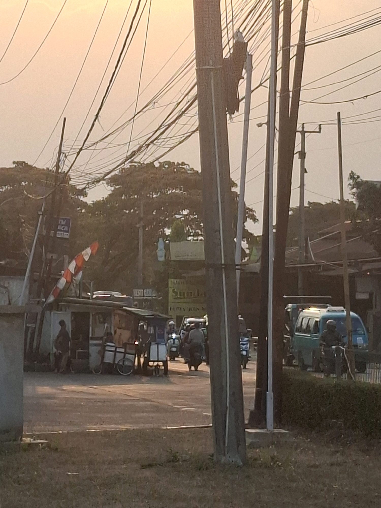 The image depicts a street scene in Karangtengah, Indonesia, characterized by a high density of overhead electrical wires. In the foreground, a grassy area is visible, leading to a paved road. Several utility poles, some appearing weathered and aged, dominate the scene, supporting a chaotic network of wires.
On the left side of the road, there is a small, open-air structure with people standing and a cart nearby. A red and white striped flag or banner is attached to a pole above this structure. Further down the road, a blue minivan is parked, with a person on a motorcycle approaching it. Several other motorcycles and a light blue van are also present, suggesting traffic.
In the background, trees and buildings are partially obscured by the overhead wires and the lighting conditions, which suggest it is late afternoon or early evening with warm, diffused light. A sign with Indonesian text, possibly advertising, is visible on a building. The overall impression is one of a busy, functional urban environment with extensive electrical infrastructure.