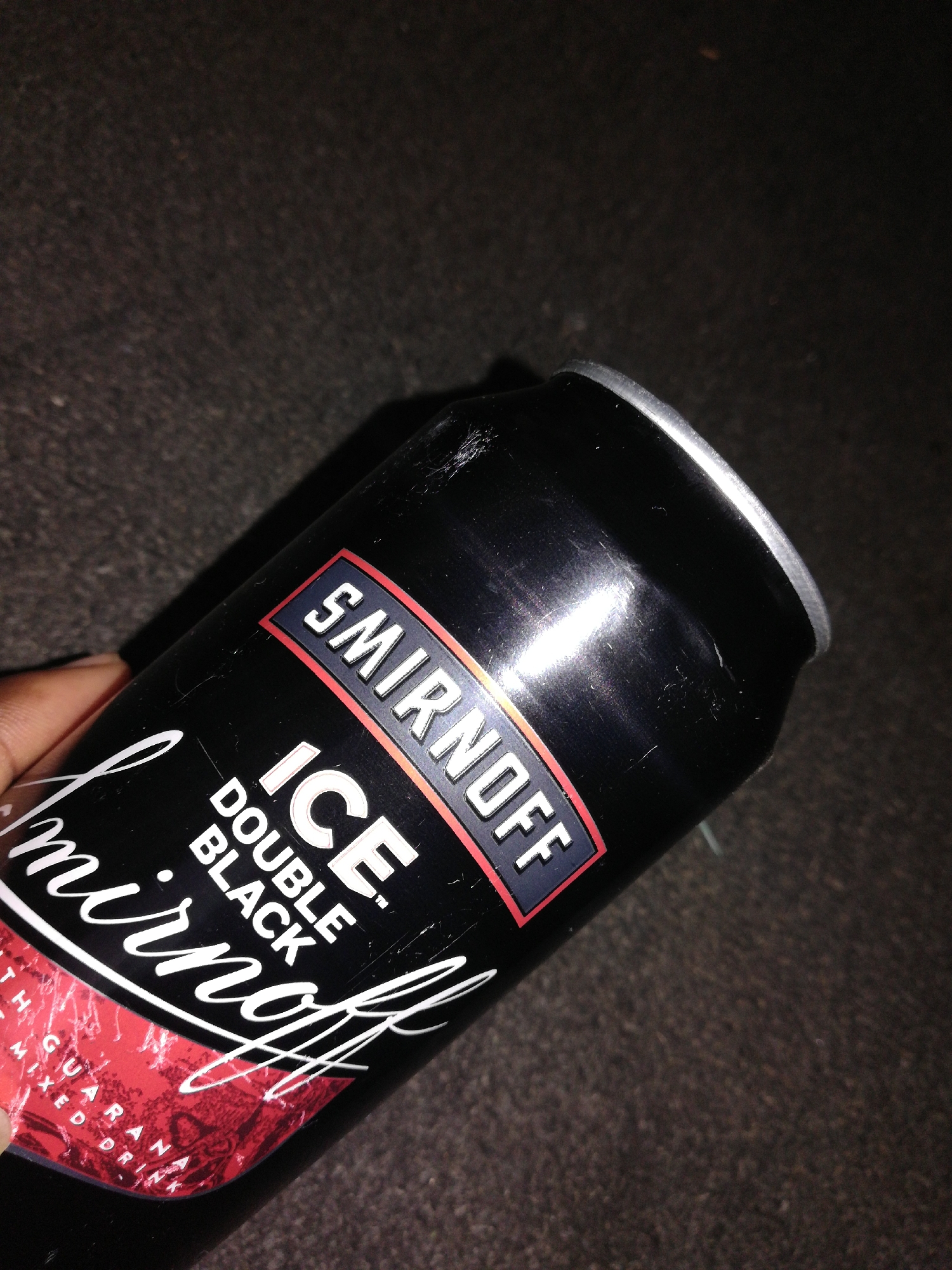 A close-up shot depicts a black can of Smirnoff Ice Double Black. The can is held at an angle, showing the top of the can, where the opening is located. The Smirnoff logo and "ICE DOUBLE BLACK" are visible on the side of the can. A hand is seen partially holding the can. The environment appears to be a dark, possibly carpeted, surface. The location is Unknown, Unknown.