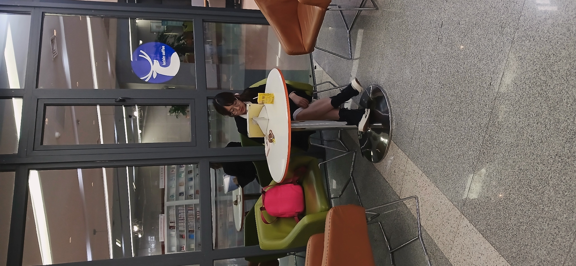 The scene is inside a Luckin Coffee location, inferred from the logo visible through the glass window. A young woman with dark hair and glasses is seated at a round table, appearing to be using a tablet. She is wearing dark socks and what appears to be casual attire. A small juice box sits on the table next to her.
Below her, a pink backpack rests on a green chair. Other chairs, upholstered in orange, are also present. The floor is tiled with a gray speckled pattern. Outside the window, a blurred interior view suggests shelves and other commercial displays.
The location is likely in the High-tech Zone of Chongqing, China, as indicated in the prompt.