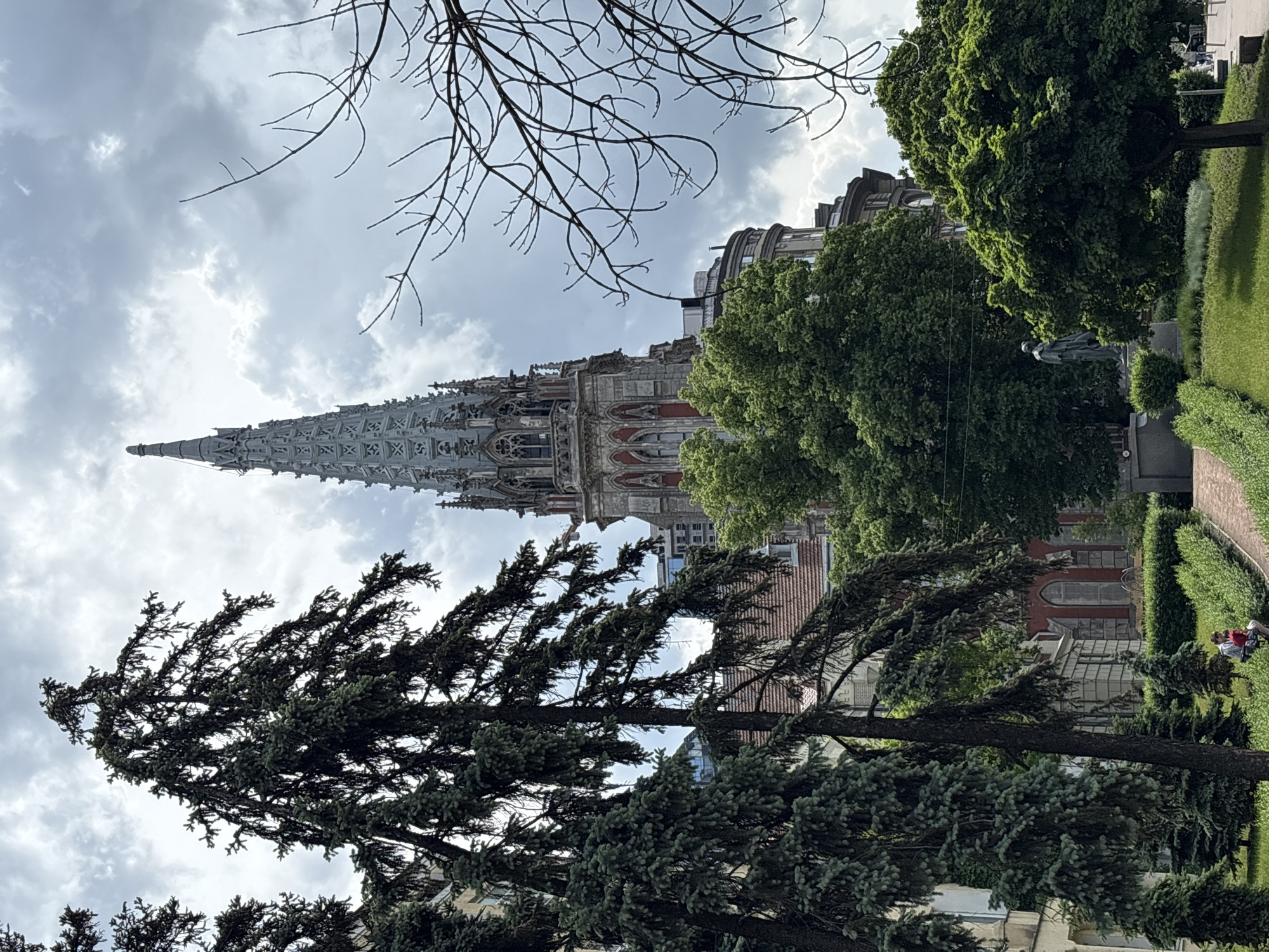 The image shows St. Nicholas Roman Catholic Cathedral in Kyiv, Ukraine, framed by trees and a cloudy sky. The cathedral's ornate spire is the focal point, rising above the surrounding greenery. A statue stands in a park-like setting near the cathedral, and people sit on a bench along a pathway. The perspective is from a slightly lower angle, looking up at the cathedral. Bare tree branches are visible in the upper left corner.