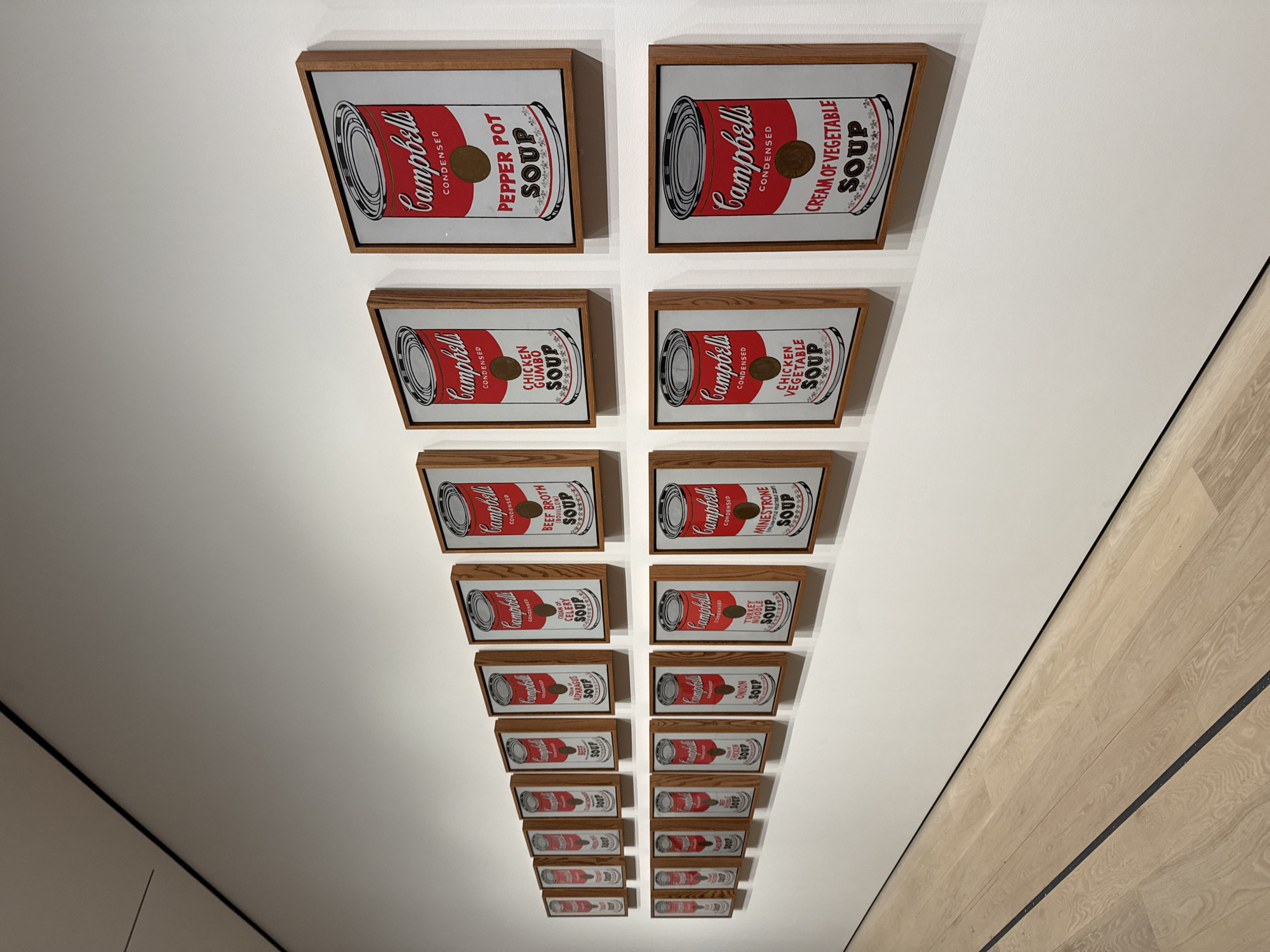 The image captures an art installation featuring multiple framed prints of Andy Warhol's iconic Campbell's Soup Cans. The prints are displayed on a white wall, arranged in a converging perspective, with larger prints at the top and progressively smaller ones towards the bottom, creating an illusion of depth. The frames appear to be made of wood. The soup can prints depict various flavors of Campbell's soup, like Pepper Pot, Cream of Vegetable, Chicken Gumbo, and Beef Broth. The right side of the frame is along the wooden floor, implying that the camera is angled upward. This installation is likely located within an art gallery or museum in New York City, where such artwork is commonly exhibited.