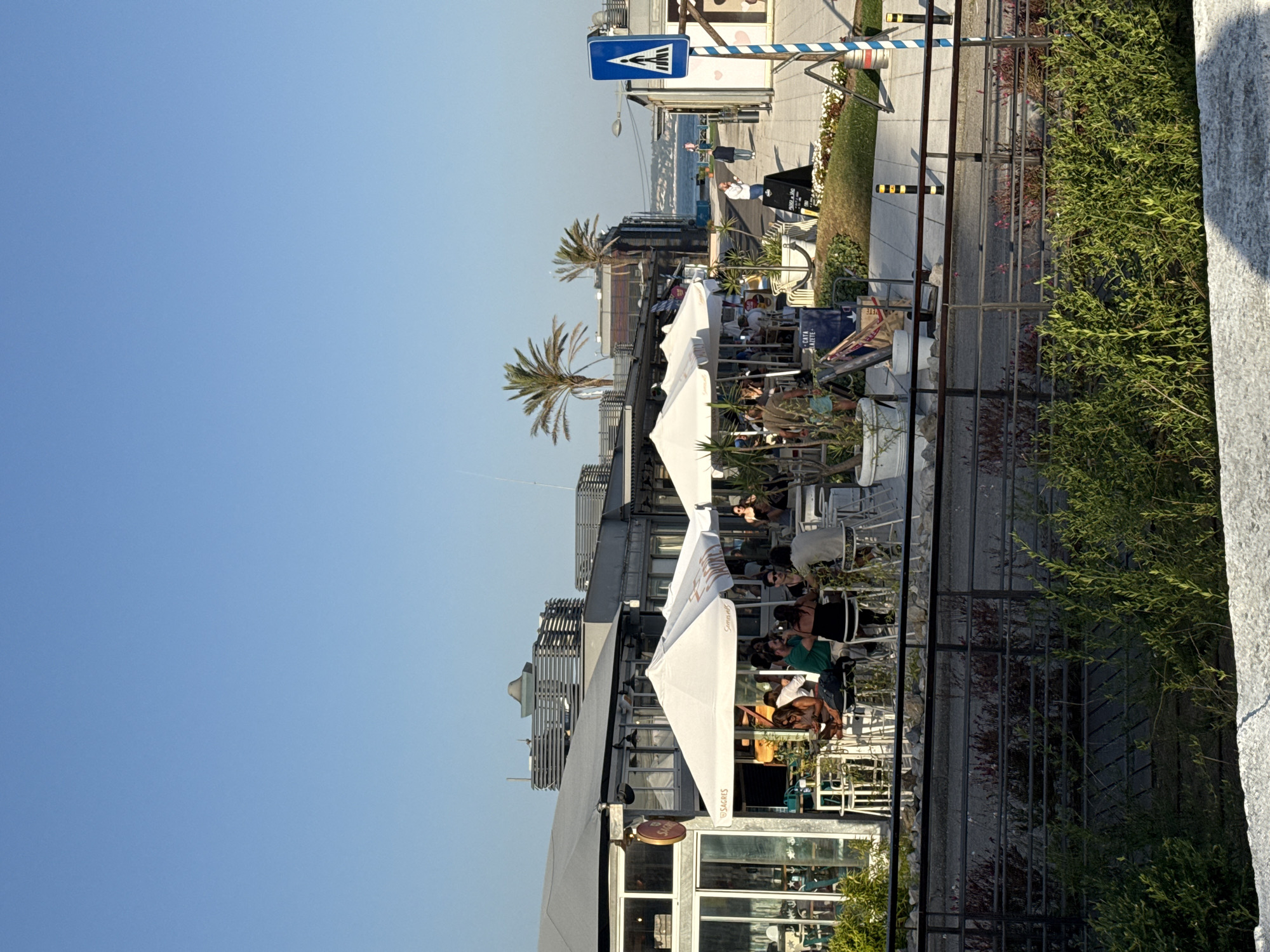 The image depicts an outdoor dining area with several tables and patrons seated under white umbrellas. The scene is situated in Oeiras, Portugal, with a view of the Atlantic Ocean in the background. The architecture suggests a modern design, with sleek buildings and metallic accents. Palm trees are visible, adding to the coastal ambiance. A pedestrian crossing sign and bollards indicate a walkway along the waterfront. People are engaged in conversation and enjoying their meals, suggesting a relaxed atmosphere. The overall impression is one of a vibrant and picturesque seaside dining establishment.
