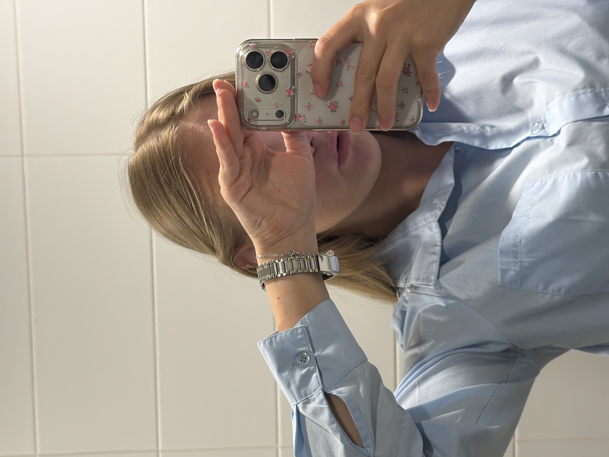 A person with blond hair, wearing a light blue button-down shirt and a metal watch, is taking a selfie in a room with white tiled walls. The phone has a transparent case with a floral pattern. The location cannot be determined based on the image alone.