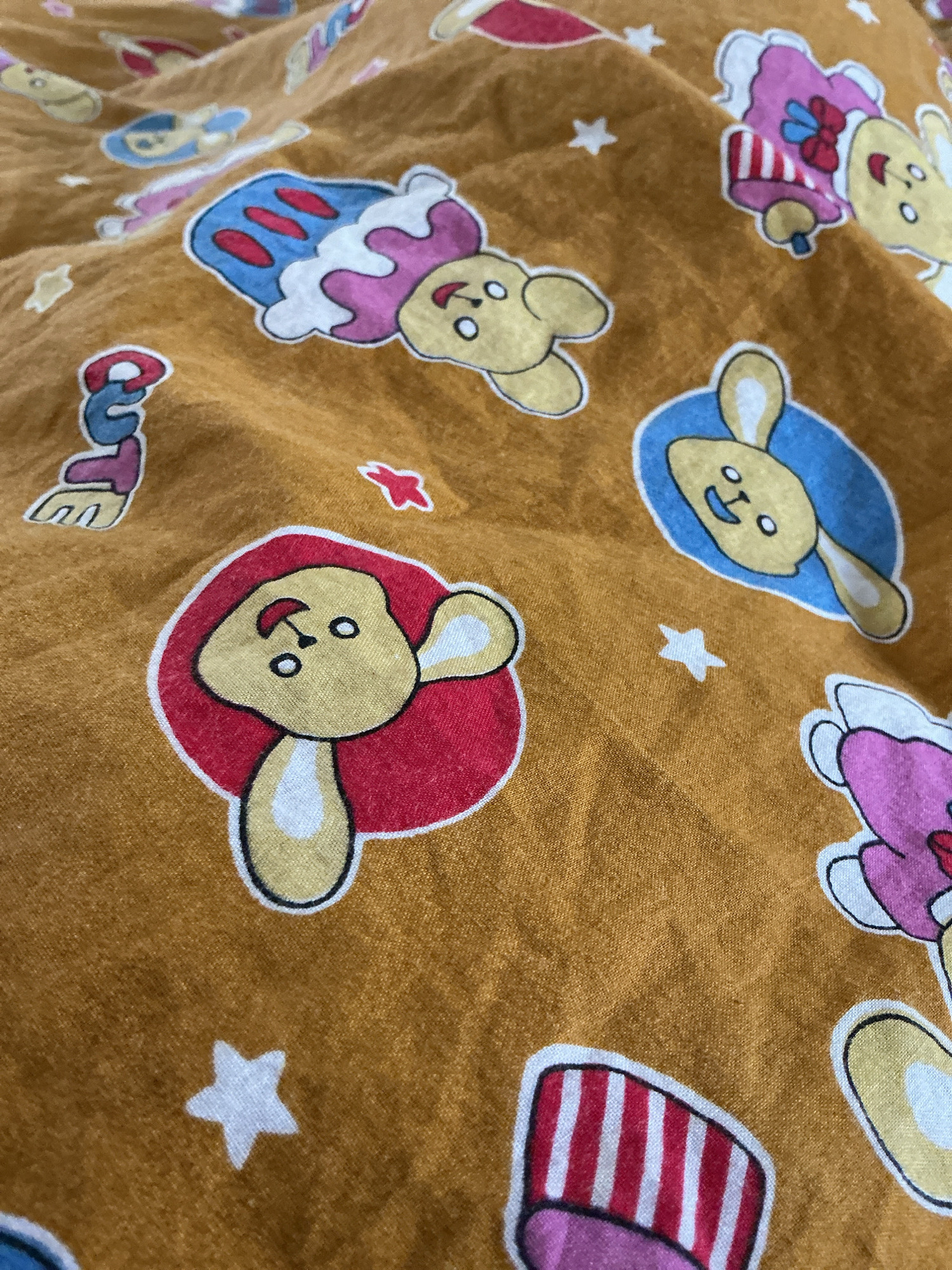 The image displays a close-up, overhead view of a brown fabric adorned with a repeating pattern of cartoon-style characters. These characters, primarily depicted as yellow, anthropomorphic animals with large eyes, appear in various poses and attire, including what looks like a blue and white dress and a pink outfit with a bow. Some characters are enclosed in circular or oval frames of red or blue. Small white stars are scattered across the fabric. The fabric itself appears somewhat creased, suggesting it might be part of bedding or clothing. The scene is a detailed, forensic description of the fabric's pattern and texture. The location in the city of Vanasthali, India, is not depicted or inferable from the visual information provided in the image.