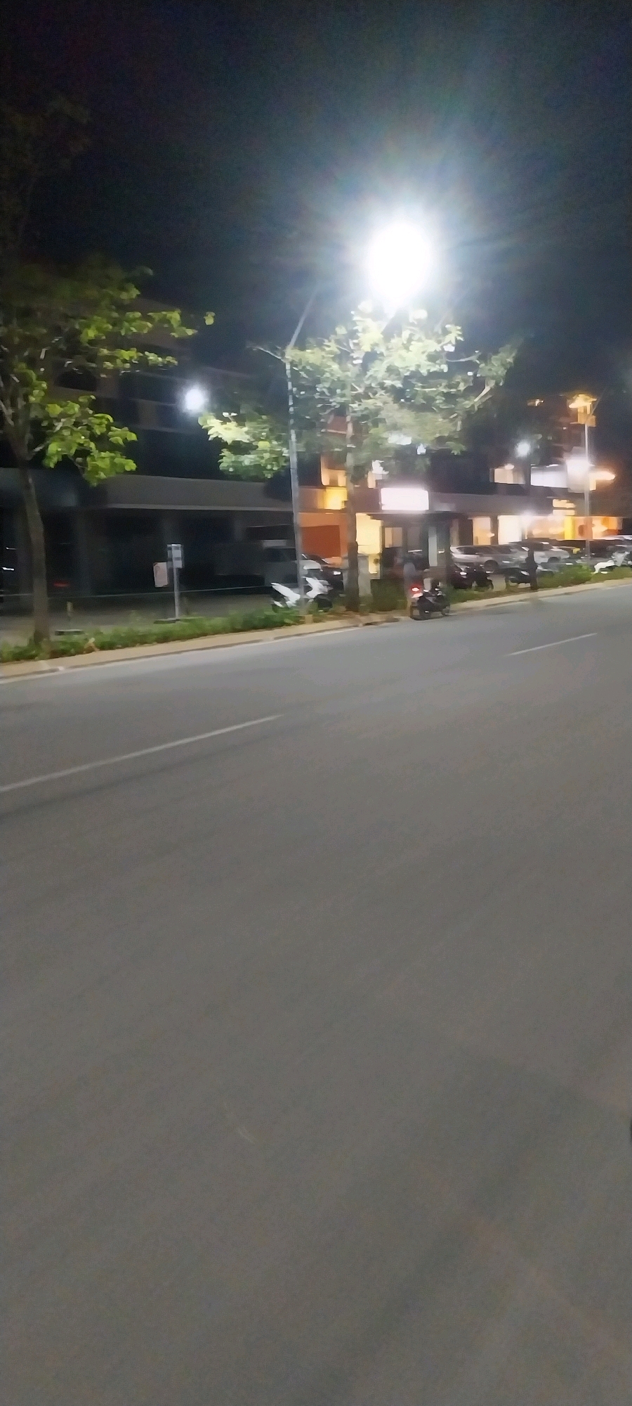 The image depicts a nighttime scene on a street in the city of Medan, Indonesia. The view is from a moving vehicle, likely a car or motorcycle, looking towards the right side of the road. The street is paved with asphalt and has a white lane marking visible. On the right side of the street, there is a sidewalk area lined with green foliage and trees. Beyond the sidewalk, a building with multiple floors is visible, illuminated by artificial lights. Several vehicles, including cars and motorcycles, are parked along the street in front of the building. A bright street lamp is positioned above the sidewalk and trees, casting a strong light. The overall environment suggests an urban setting at night, with the presence of commercial or residential buildings and street lighting. There are no clearly identifiable people performing specific actions, but the parked vehicles imply activity in the area.