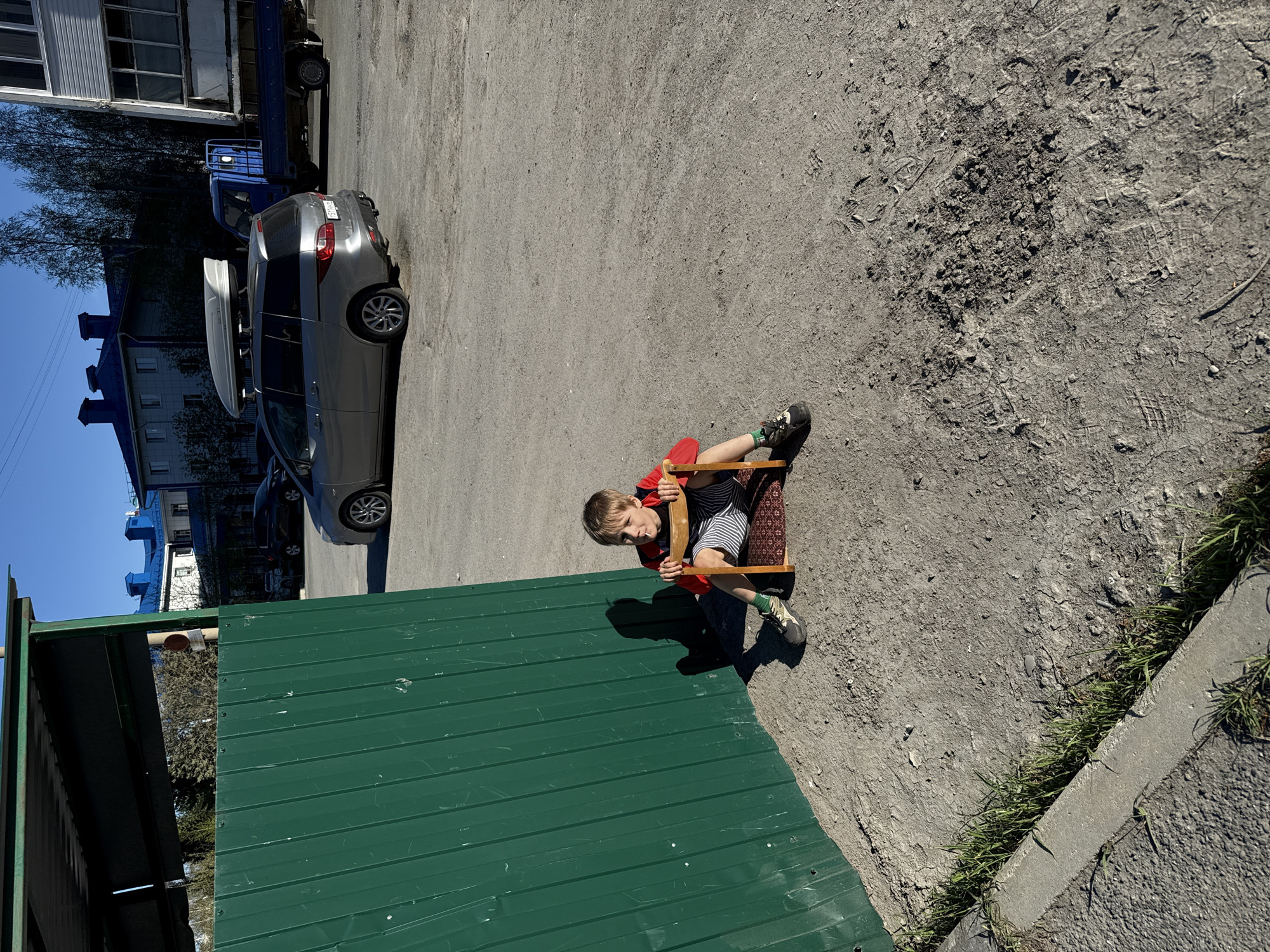 The image shows a child sitting on a wooden chair in what appears to be an urban outdoor area. The child is wearing a red shirt and striped shorts. The ground is a mix of asphalt and dirt with some vegetation visible. A green metal structure is in the lower left corner of the image. In the background, there is a gray car, a blue truck, and buildings, suggesting the location is in the city of Tobolsk, Russia.