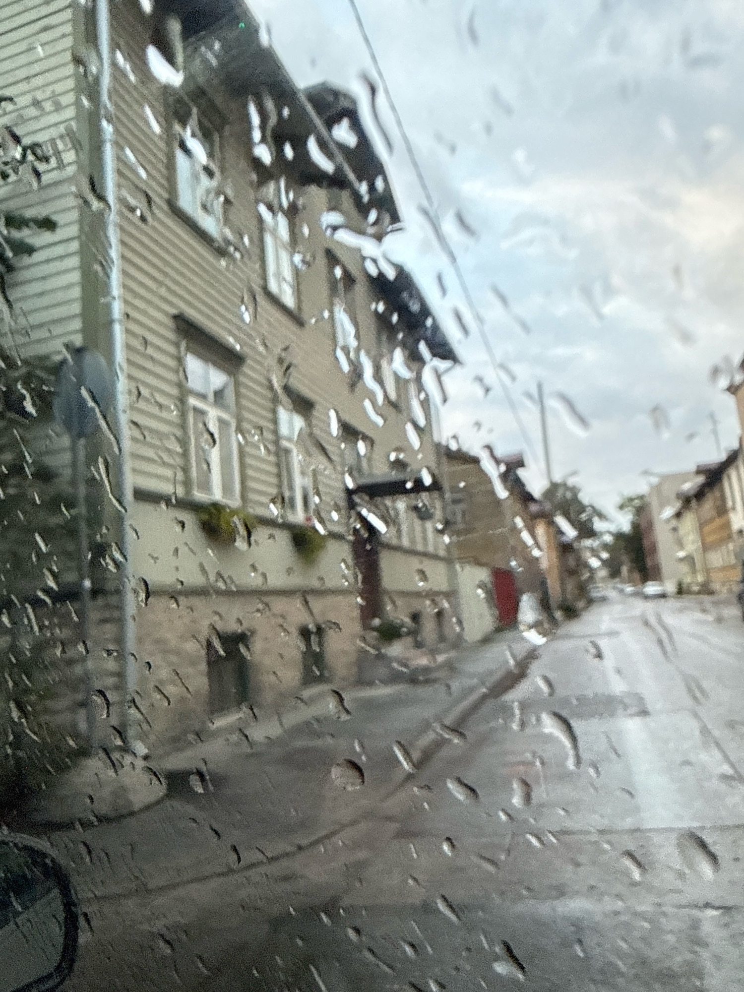 This is a photograph taken through a rain-streaked window, likely from inside a vehicle, looking out onto a street in Tallinn, Estonia. The foreground is dominated by water droplets on the glass, distorting the view of the scene beyond.
The primary subject is a row of buildings, appearing to be residential or mixed-use, with a distinct architectural style characterized by wooden siding and pitched roofs. The building closest to the viewer on the left is light-colored, possibly beige or light gray, with horizontal wooden planks. It has several windows, some with visible panes, and a lower section that appears to be made of stone or brick. Planter boxes with greenery are visible on a ledge beneath some of the upper windows.
Further down the street, other buildings are visible, some with darker facades, including one with a red door. The street itself is wet and appears to be paved, with reflections suggesting it is a city street. The overall atmosphere is overcast and damp, consistent with rainy weather. The sky is cloudy and muted.
The perspective suggests the viewer is looking from a street level or slightly elevated position. No people are clearly discernible. The image captures a slice of urban life in Tallinn during a period of rain.