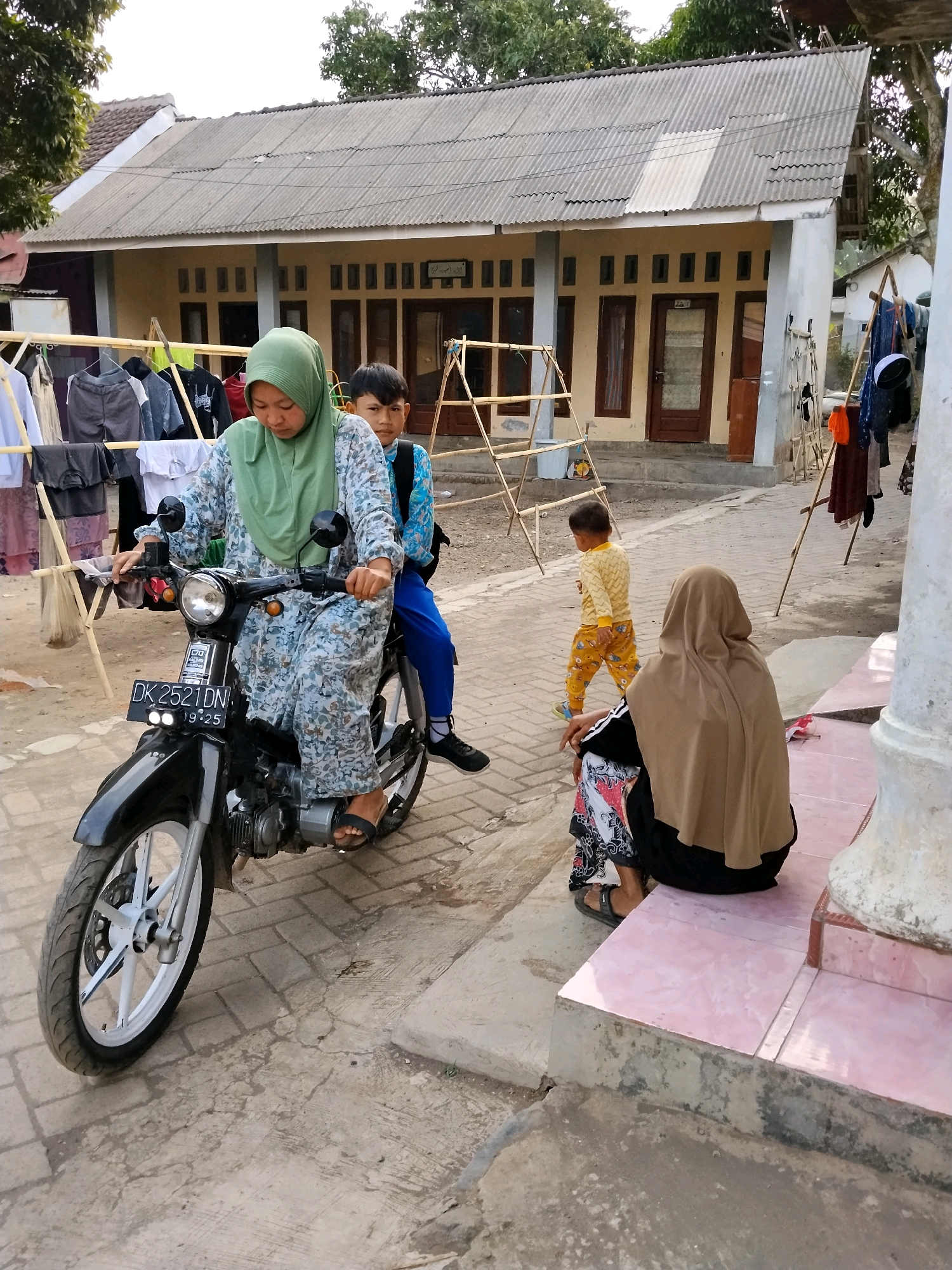 In Panggungan, Indonesia, a woman wearing a green hijab and a patterned dress rides a black motorcycle with a young boy in a blue shirt and backpack as a passenger. A toddler in a yellow shirt and orange pants walks away from the motorcycle towards the right side of the frame. Another person wearing a brown hijab and a patterned skirt is seated on pink tiles to the right. In the background, a house with a corrugated metal roof is visible, with clothes drying on a rack in front of it. The scene appears to be a residential street.