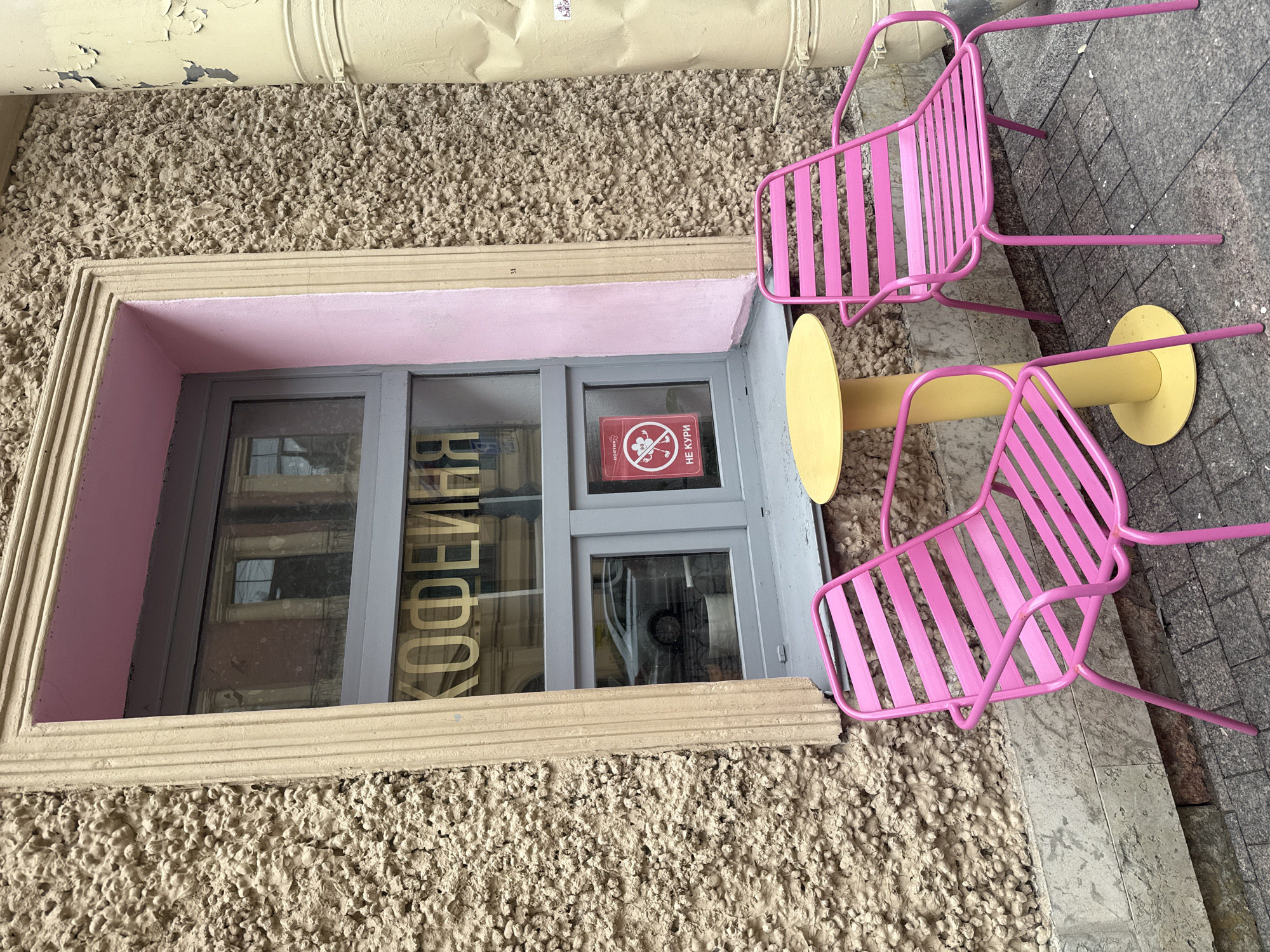 An outdoor scene captures the entrance of a coffee shop, featuring a distinctive building facade with a vibrant, modern seating arrangement. The image is primarily composed of a roughly textured, stucco-like wall in muted beige, adorned with a light beige window frame. Above the window, the wall is painted a soft pink.
The focal point is a large window with a grey frame, reflecting what appears to be other buildings across the street. Across the window in large, gold letters, the word "КОФЕЙНЯ" (KOFEYNYA), meaning "Coffee Shop" in Russian, is visible, indicating the establishment's purpose. Inside the window, a red sign with a white circle and a crossed-out cigarette icon clearly states "НЕ КУРИ" (NE KURI), meaning "No Smoking."
In front of the window, two bright pink metal slatted chairs and a single round yellow metal table are arranged, suggesting an outdoor seating area for patrons. The chairs and table are placed on a tiled or paved ground surface, distinct from the textured wall. Above the scene, a large, yellow pipe with peeling paint runs horizontally, adding a touch of urban decay to the otherwise colorful and inviting setup. The overall impression is that of a charming, slightly eclectic coffee shop entrance on a street, likely in Saint Petersburg, Russia, during daytime hours. No people are visible.