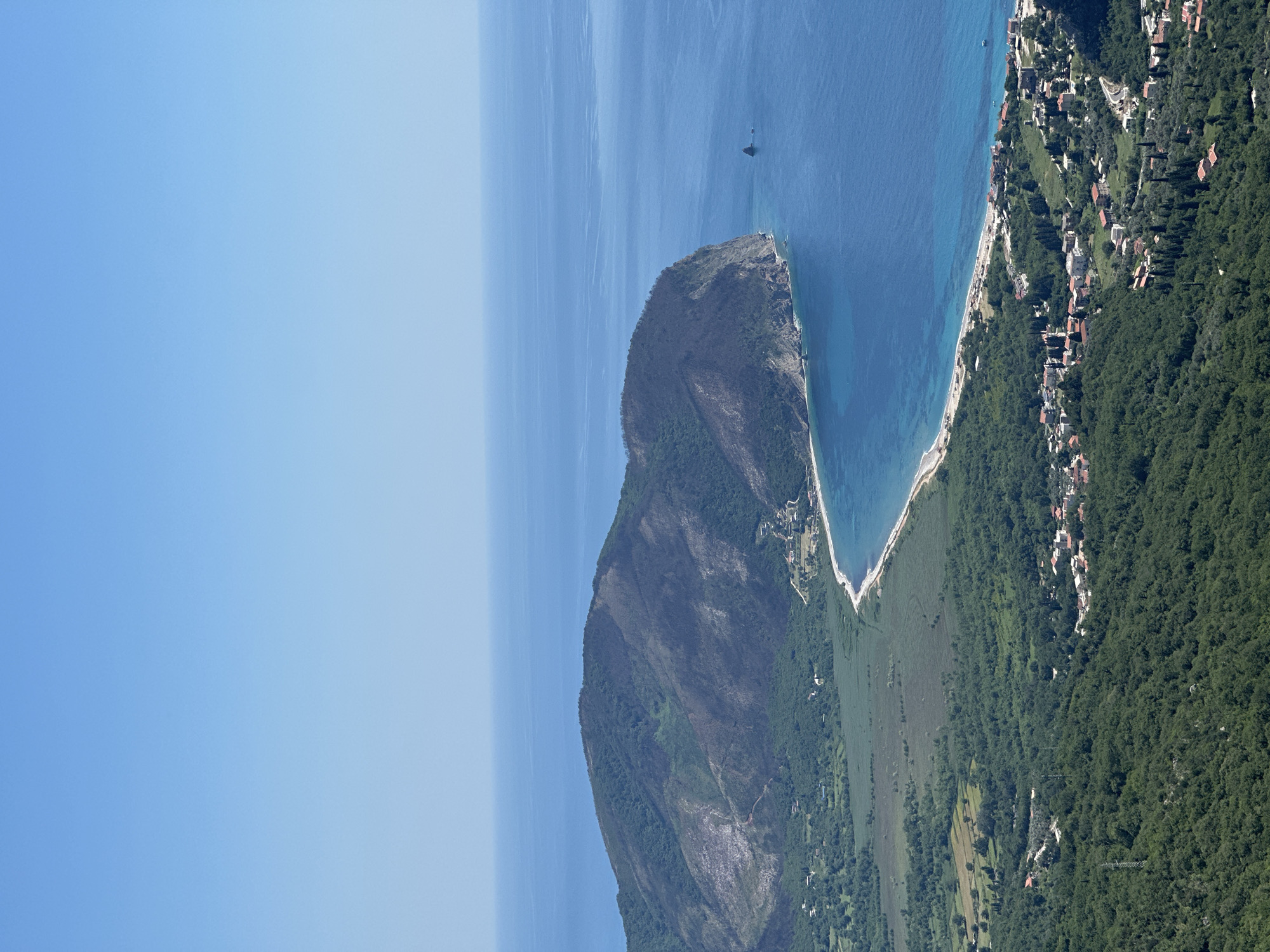 The image shows a coastal landscape in Novoselje, Montenegro. The scene includes a large, predominantly barren mountain sloping down to a narrow coastal plain and a crescent-shaped beach. The Adriatic Sea borders the mountain and beach, with a boat visible in the distance. Adjacent to the beach is a settlement with houses nestled within dense, green vegetation. The sky is a clear, cloudless blue.