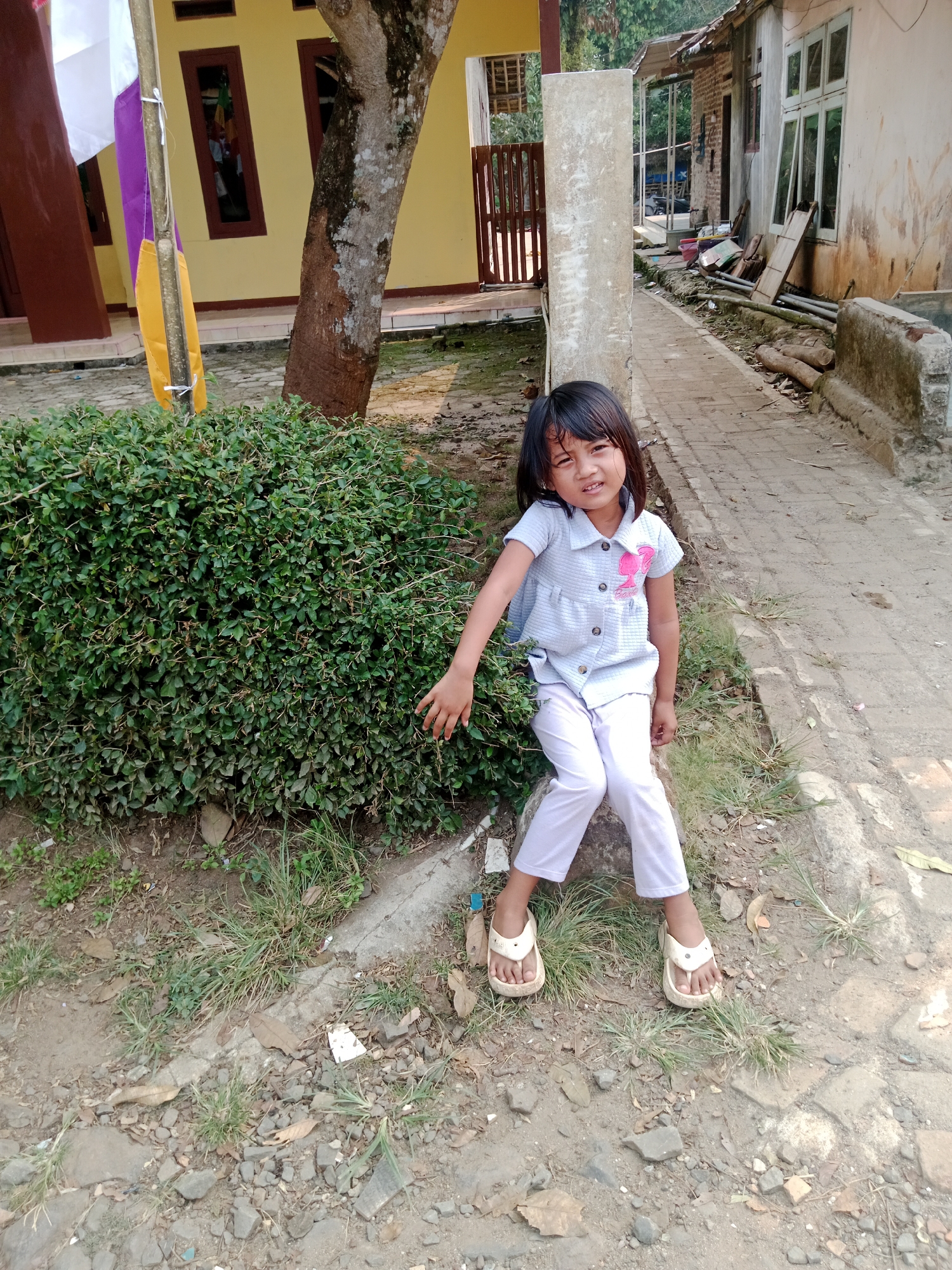 A young Indonesian girl with dark hair and bangs, wearing a light blue patterned shirt with a pink embellishment and white pants, sits on a stone by a dense green bush. Her feet, adorned with beige sandals, rest on the ground. To her left, a tree trunk is visible, and a flag pole with white, purple, and yellow flags stands nearby. Behind her, a yellow building with brown-trimmed windows and a wooden fence can be seen. To her right, a brick pathway extends into the background, flanked by a weathered building and debris. The ground is a mix of dirt, rocks, and dry leaves.