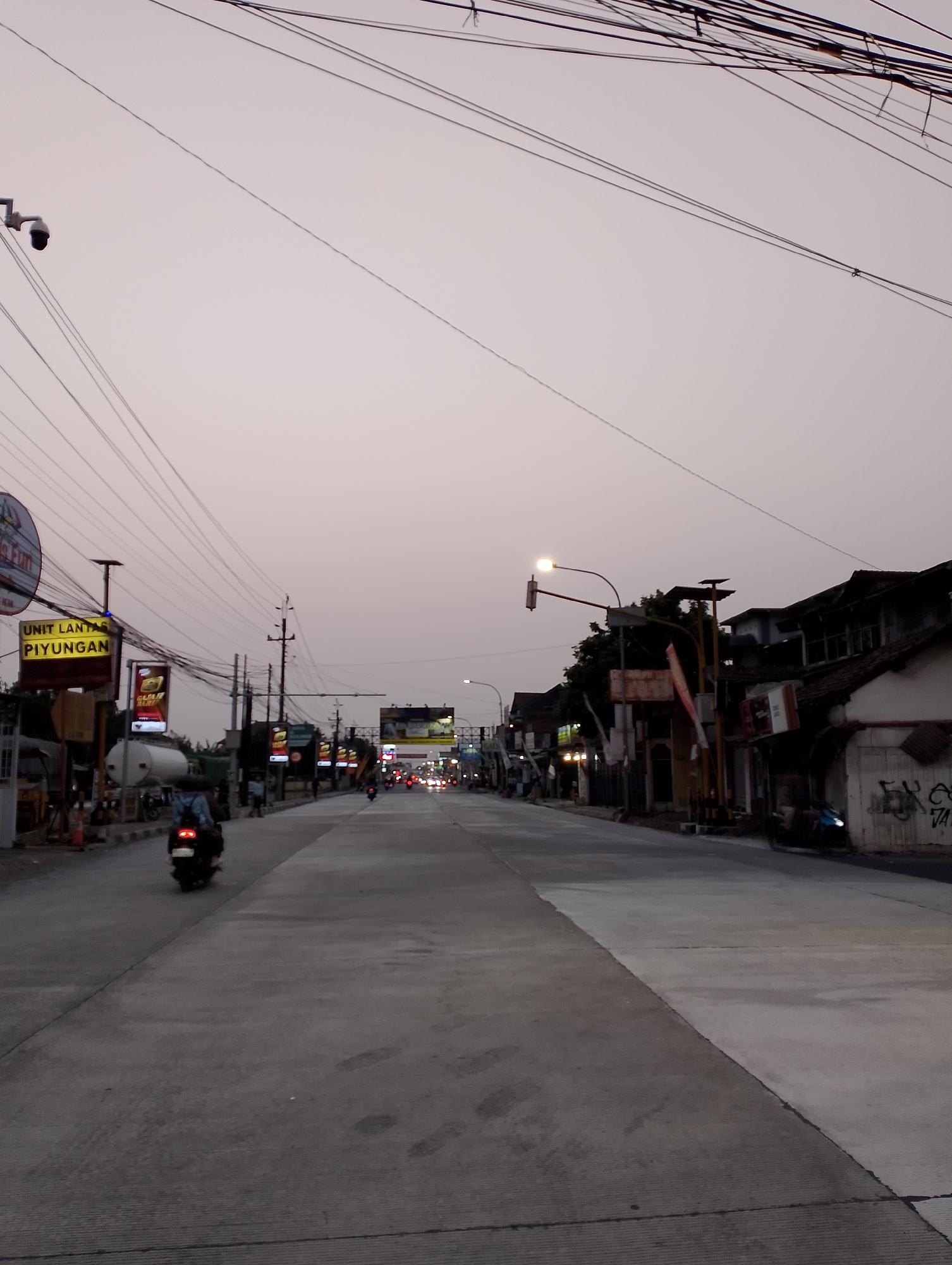 This is a medium-shot, eye-level, vertical photograph of a street scene in Srimartani, Indonesia, during twilight. The sky is a pale, overcast gray. The street is paved with concrete and has visible expansion joints. On the left side of the street, there are several businesses with signs, including one that reads "UNIT LANTAS PIYUNGAN". A white tanker truck is parked to the left of the road, and traffic cones are visible. A person on a scooter, wearing a backpack, is riding away from the camera on the left side of the street. Further down the street, a line of streetlights and illuminated signs are visible, with traffic moving in the distance, indicated by the glow of headlights and taillights. Buildings line the right side of the street, some with graffiti on their walls. Overhead, a dense network of electrical wires stretches across the frame. The overall impression is of a quiet, somewhat industrial or commercial area as evening settles.
