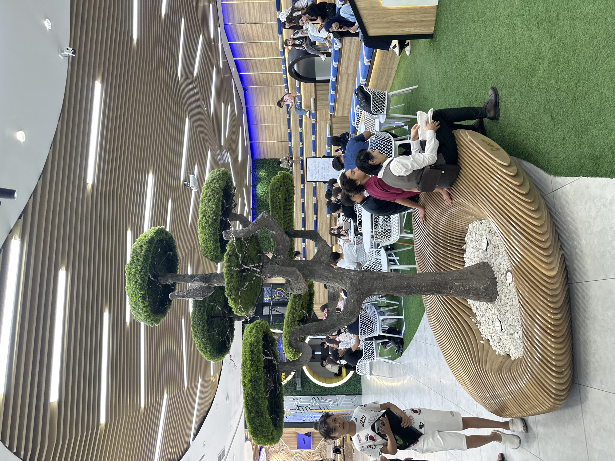 The image depicts an indoor space in Tashkent, Uzbekistan, possibly a library or study area. The environment is characterized by a high ceiling with linear light fixtures and a wood-paneled design. A large artificial tree with rounded green foliage dominates the foreground. The tree is integrated into a circular, tiered wooden seating structure filled with white pebbles at its base. Several people are present in the scene. Some are seated at tables and chairs further back, engaging in activities like reading or studying. A person wearing dark trousers and brown shoes sits on the wooden structure, also engrossed in reading a book. Near the bottom, a child is standing, holding a black object. The space includes artificial green turf in certain areas, adding a touch of greenery.