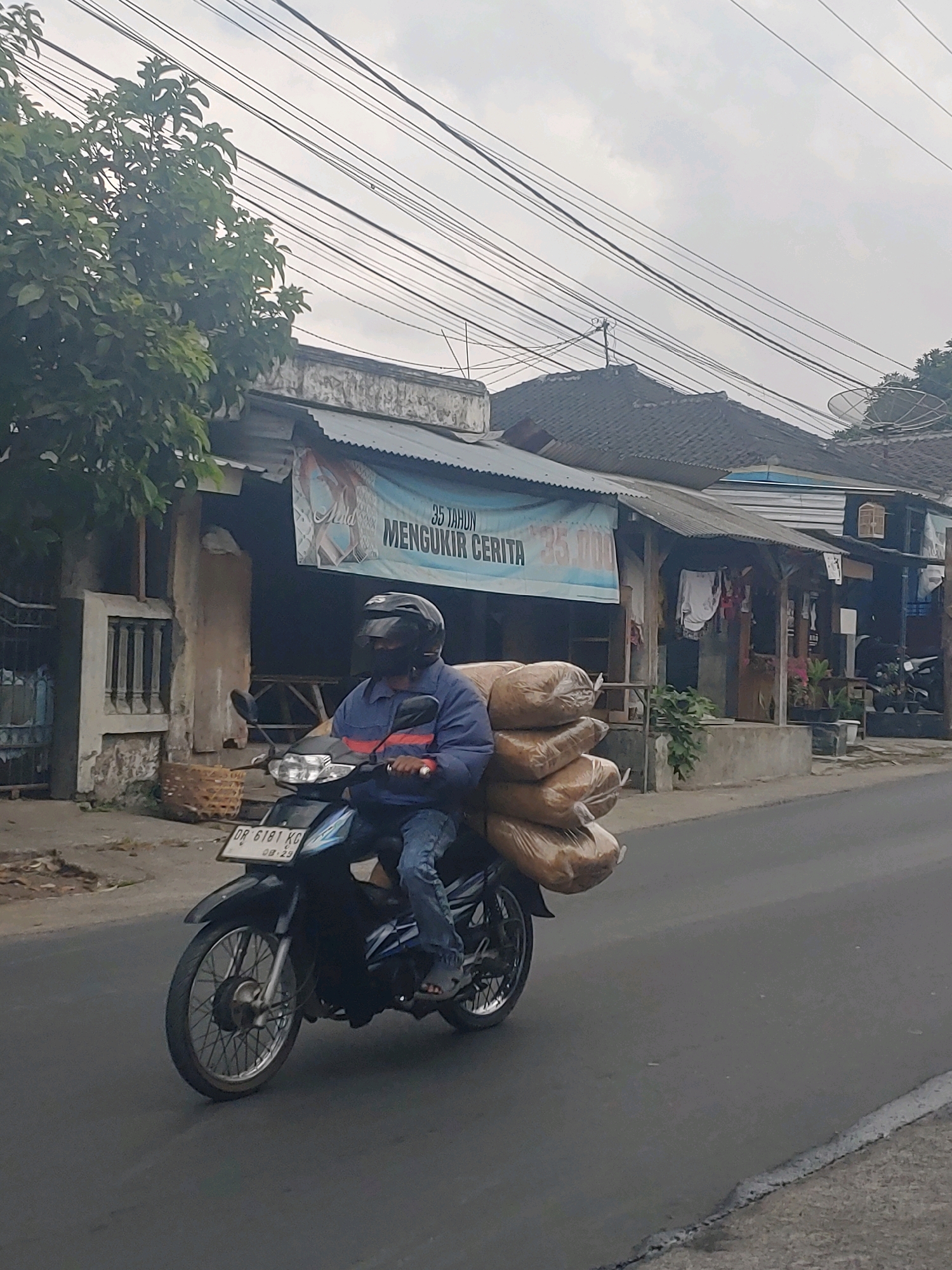 A medium shot shows a person riding a black motorcycle loaded with several large, brown, sack-like packages on a paved street in Kembang Kerang, Indonesia. The rider is wearing a dark helmet with a visor, a blue jacket with orange stripes on the sleeves, and blue jeans. The motorcycle has a blue and silver design on its side and a license plate reading "DR 6181 KG 08-29".
The background features a row of buildings with corrugated metal roofs. A banner hangs across the front of one building with Indonesian text that reads "35 TAHUN MENGUKIR CERITA 35.000". There are trees and overhead wires visible. The sky is overcast. The scene is captured from a slightly low angle, looking up at the rider and the buildings.