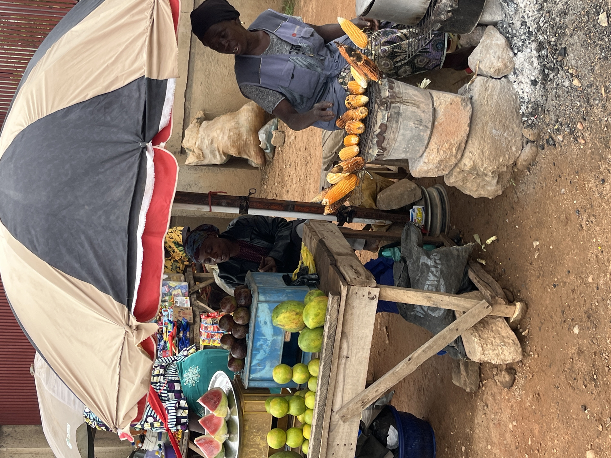 In Bukuru, Nigeria, an outdoor market scene unfolds under a large, striped umbrella. Two women are engaged in selling produce and grilled corn. On the left, a woman, partially visible, is attending to a display of sliced watermelons and other fruits. In the center and right, another woman, wearing a dark headscarf and a light blue vest over a patterned shirt, is actively grilling corn on a metal grate over coals. The corn cobs are arranged on the grate, some appearing to be fully cooked.
The market stall is constructed from rough-hewn wooden planks, with fruits like oranges and papayas displayed prominently. Behind the produce, a blue plastic crate holds dark, round fruits, possibly passion fruits. The ground is a mixture of reddish-brown dirt and scattered stones. A large, light-colored burlap sack is visible in the background, leaning against a wall. Various other market goods, including packaged snacks and other fruits, are present, suggesting a bustling retail environment. The overall impression is of a lively, informal marketplace.