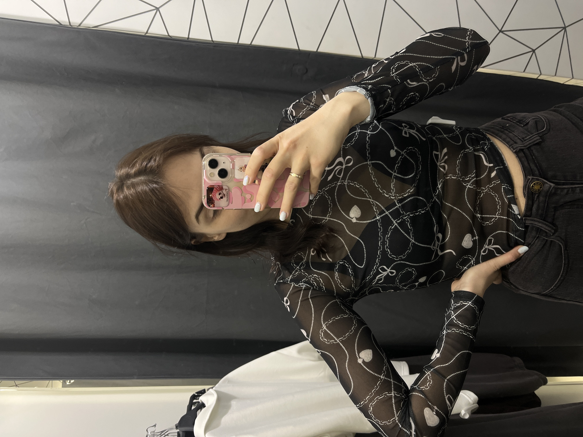 A person with brown hair is taking a selfie in a dressing room. They are holding a pink phone with a cartoon case. They are wearing a long-sleeved, sheer black top with white chain and heart patterns, and dark grey jeans. A white shirt is visible on a hanger to the left. The dressing room has a black background and a patterned black and white border at the top. The picture might be taken in the city of Shushary, Russia.