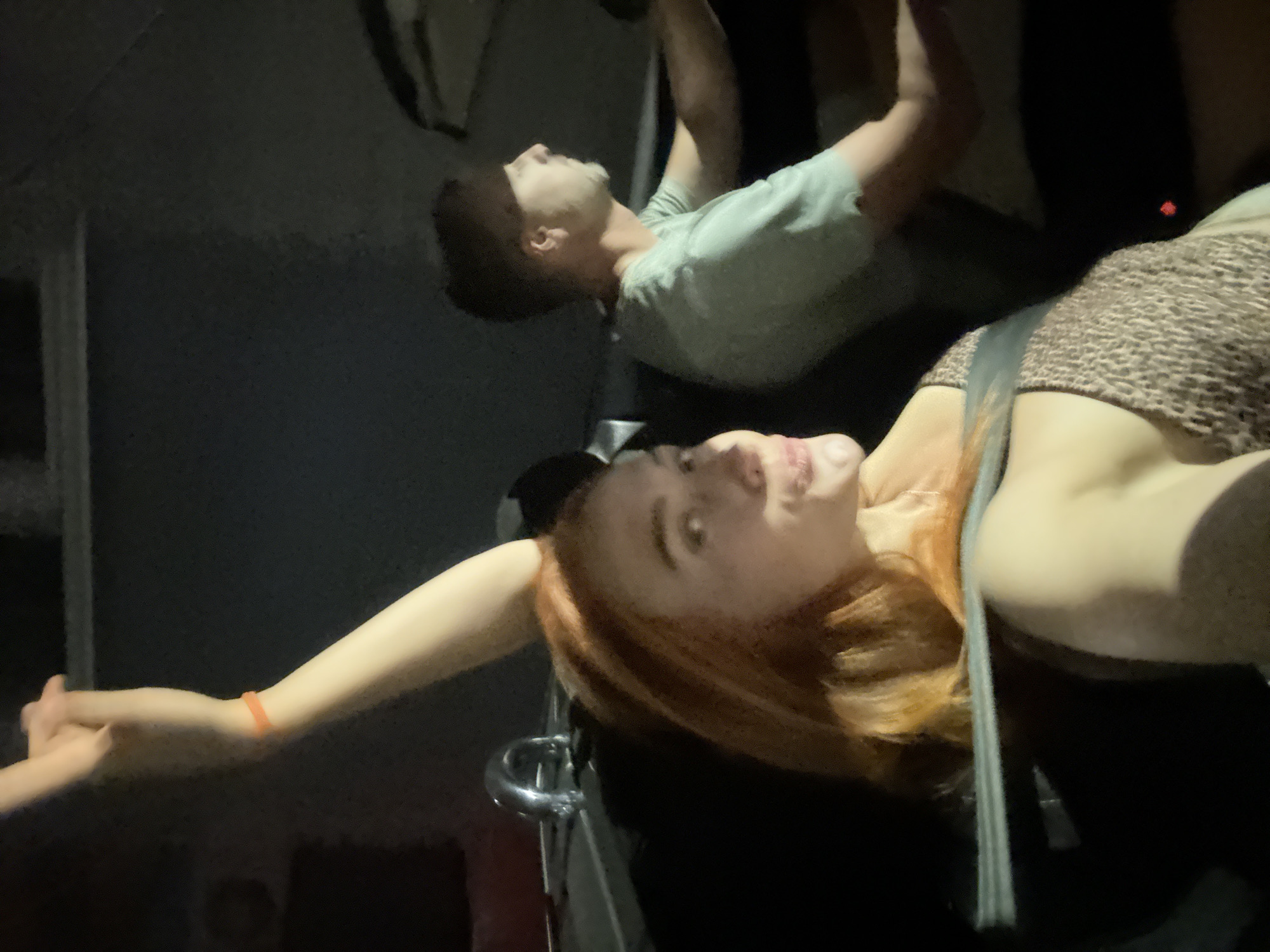 The image shows two individuals in a vehicle at night, possibly in Budva, Montenegro. The person in the foreground, a woman with red hair, is looking towards the camera with her left arm raised and her hand making a gesture. She is wearing a leopard-print top and an orange bracelet on her left wrist. A seatbelt is visible across her chest. Behind her, a man with short brown hair and a goatee is driving the vehicle, wearing a light green t-shirt. The lighting suggests it is nighttime, and the background is dark and indistinct. The interior of the car is visible, including a metallic handle.