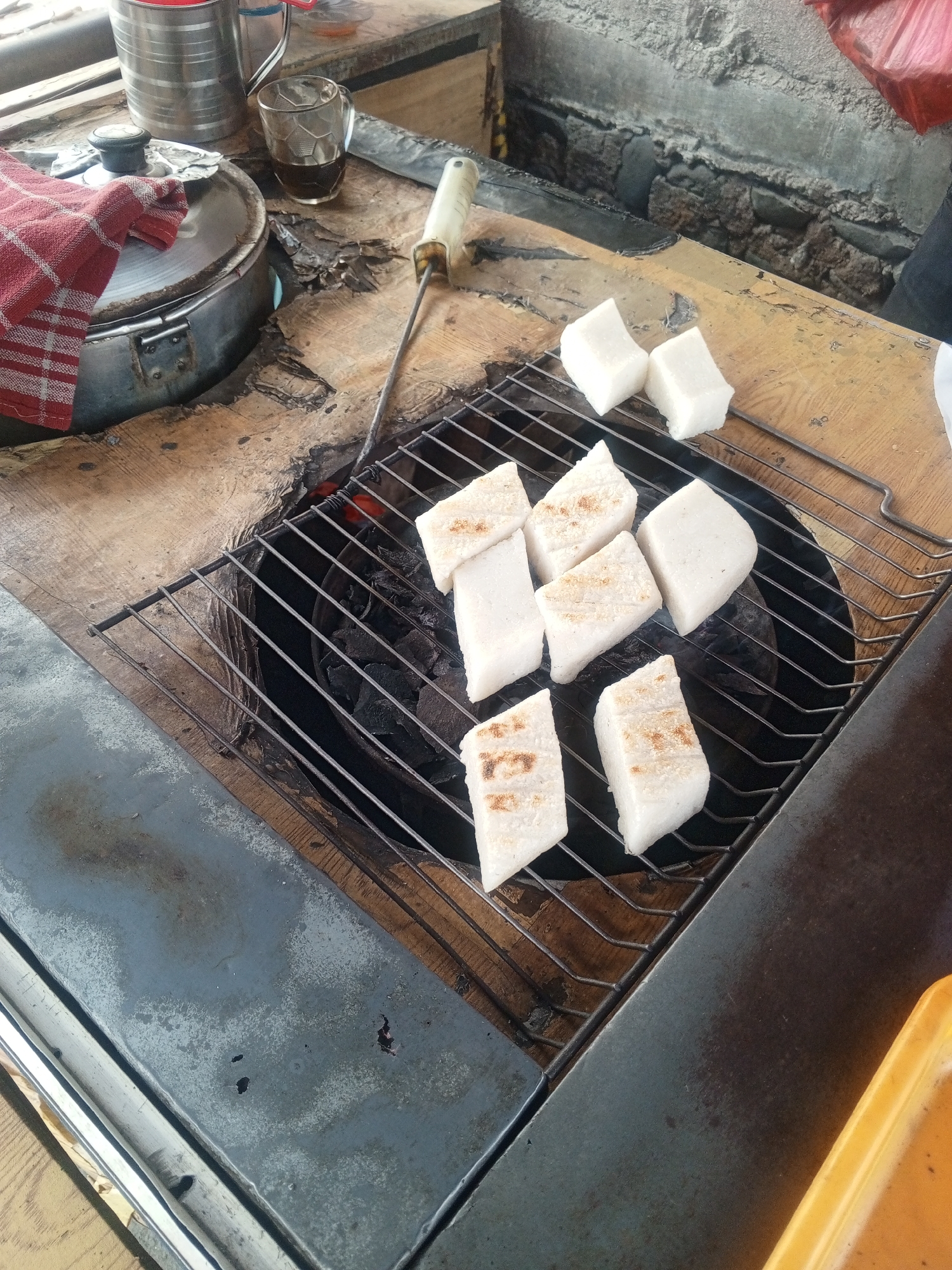 The image depicts a street food stall in Depok, Indonesia, where white, rectangular pieces of food, likely rice cakes or sticky rice, are being grilled over charcoal. The food is arranged on a metal grate above a bed of glowing coals. Some of the pieces show slight browning from the grilling process. In the background, a metal pot with a red-and-white checkered cloth draped over it sits on a wooden surface, alongside a glass of dark liquid, possibly coffee or tea. A metal skewer with a white handle is also visible. The overall environment appears to be an outdoor setting, consistent with a street food stall.