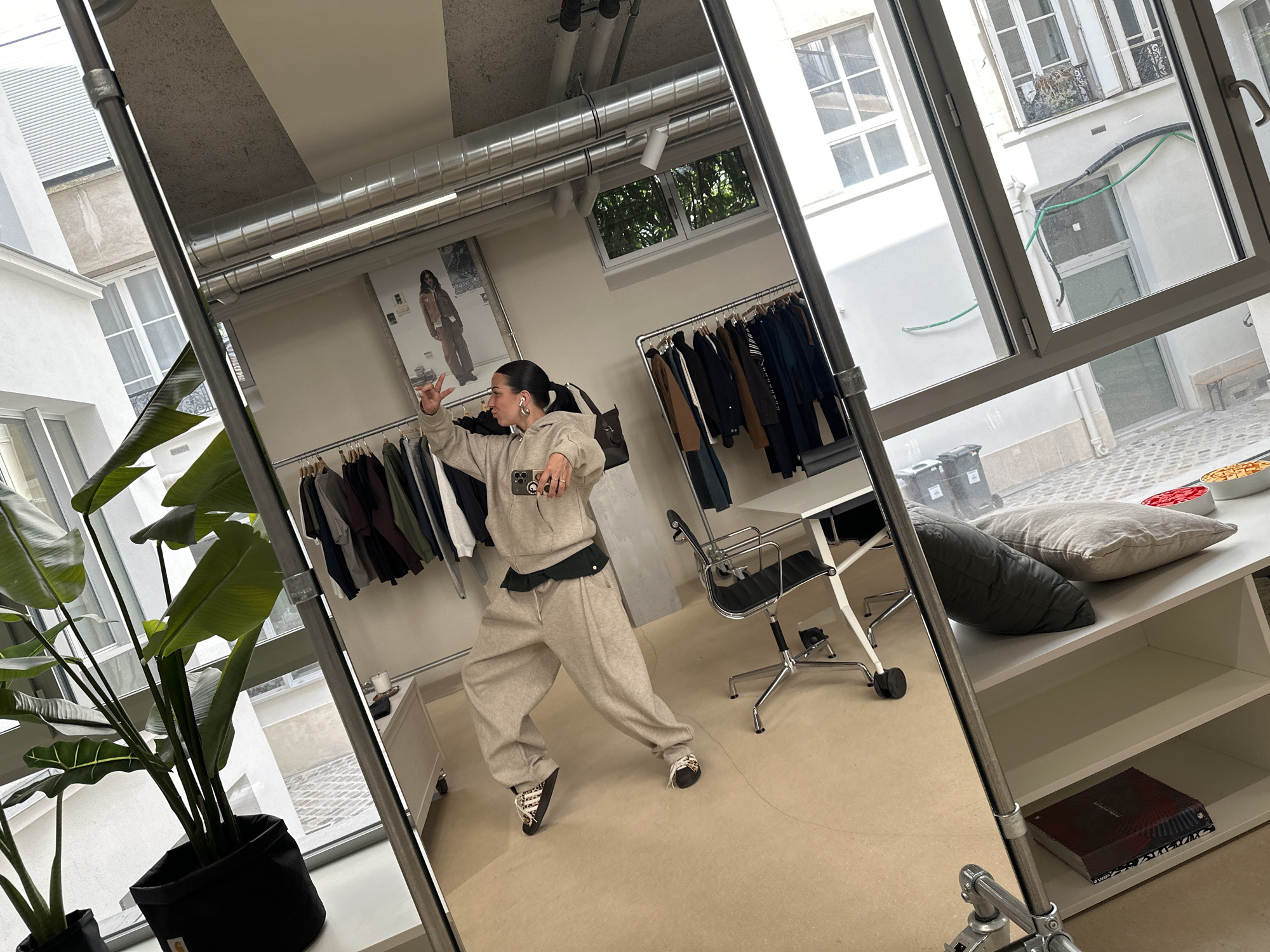 The image depicts a reflection in a large mirror. The reflection shows a person dressed in a beige tracksuit, posed as if dancing or gesturing, while holding a smartphone. Behind the person are clothing racks filled with garments, a desk with a chair, and a poster featuring a person in similar attire. Outside the window, visible both through the window itself and reflected in the mirror, are elements of a typical Parisian courtyard: white buildings, small balconies with wrought iron details, and glimpses of foliage. This suggests an interior space of a clothing store or showroom in Paris, France. The foreground of the image includes a large potted plant and a white shelf unit, further indicating an interior setting.