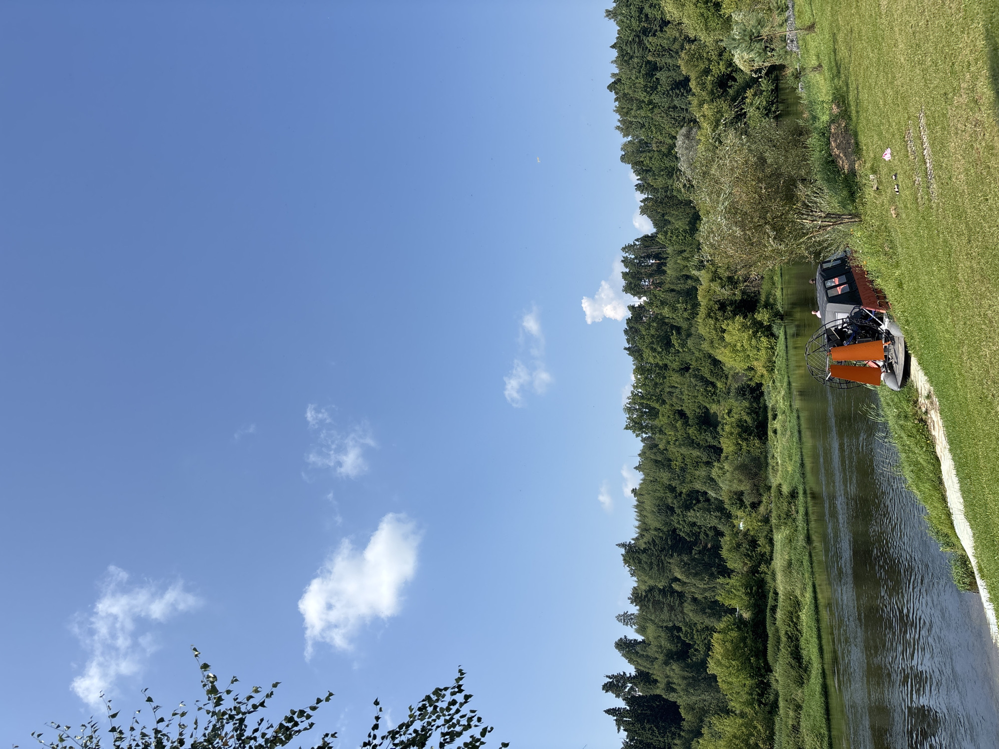 The image captures a serene riverside scene in Zvenigorod, Russia. A body of water, likely a river, flows from the left towards the right of the frame, with a dense, green forest lining its opposite bank. The sky above is a clear, bright blue, dotted with a few wispy white clouds. On the right bank, a grassy slope leads down to the water's edge. Resting on the grass, partially on a paved area, is an orange and black hovercraft. Two individuals are visible near the hovercraft, appearing to be engaged in conversation or preparing for an activity. The overall impression is one of a peaceful summer day in a natural setting.