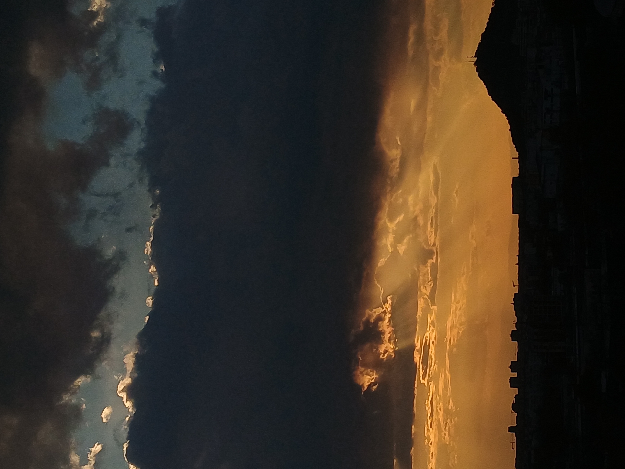 The image shows a sunset over Plovdiv, Bulgaria. The sky is split into two distinct sections. The left side is dominated by dark, heavy clouds, while the right side is illuminated by golden sunlight. A silhouette of buildings and hills is visible on the far right, suggesting an urban setting. There are no visible people, subjects, objects, actions, or interactions. The environment depicts the boundary between daylight and approaching darkness.