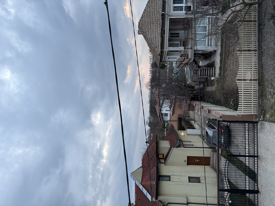 The image depicts a residential street scene in Stefan Karadja, Bulgaria, under an overcast sky with hints of orange and pink light near the horizon, suggesting either dawn or dusk. The perspective is elevated, looking down into a neighborhood.
In the immediate foreground, a dark metal gate with a decorative top design opens onto a paved driveway. To the right of the gate, a white wooden picket fence encloses a parcel of uncultivated ground. Two prominent black utility wires traverse the upper half of the image horizontally.
To the left, a two-story house with pale yellow stucco walls and a red-tiled roof features multiple gables. Its front entrance, facing the viewer, has a dark brown door adorned with a decorative wreath. A dark-colored sedan, appearing to be a BMW wagon, is parked on the driveway, positioned directly in front of this house's side entrance.
To the right, a two-story house with light grey walls and a terracotta-tiled hip roof is visible. This house features an upper-level balcony with a dark railing and an external staircase leading to a side entrance. The ground in front of this house consists of bare earth with sparse vegetation.
In the mid-ground, between the two primary houses, a narrow concrete path runs alongside a privacy wall. Bare deciduous trees and shrubs are scattered throughout the properties. The background reveals additional residential structures with various roof types, intermingled with more bare trees, extending towards a distant, slightly elevated landscape under the same cloudy sky. No individuals or discernible actions are present in the scene.