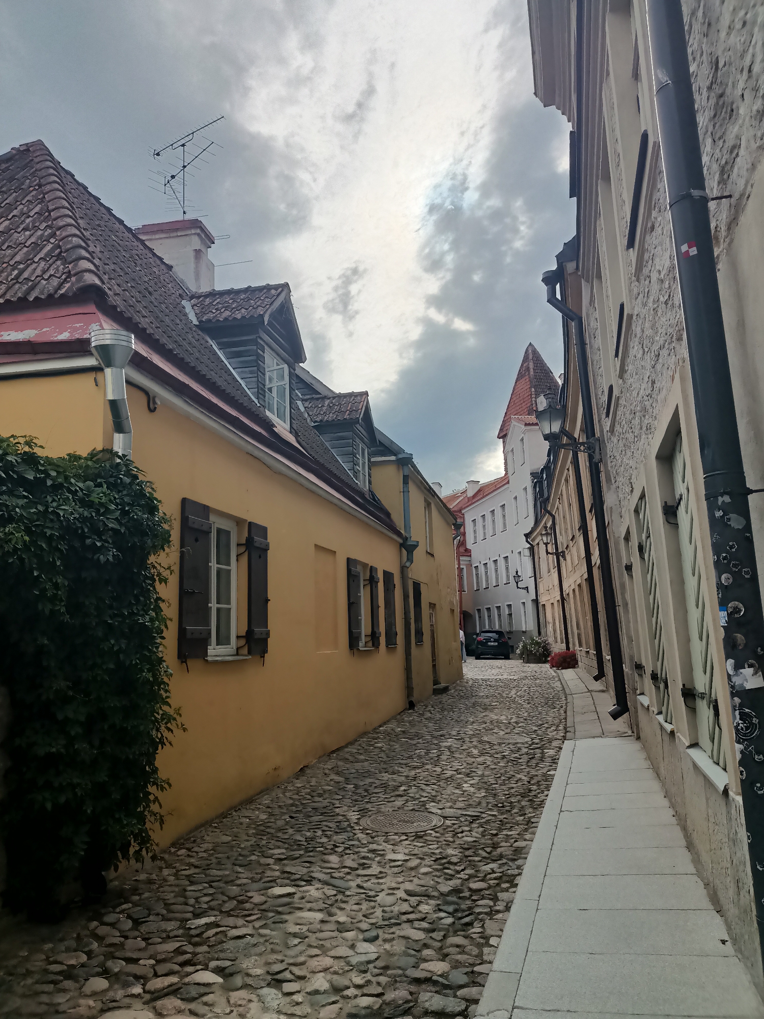 The image depicts a narrow, cobblestone street in the Old Town of Tallinn, Estonia. On the left, a building with a yellow facade features a large, dark green ivy covering its side and a visible rainwater pipe with a silver downspout. Several windows with dark wooden shutters are visible on this building. The street is paved with uneven cobblestones.
To the right of the street, a building with a stone facade has a light-colored sidewalk running alongside it. This building also has multiple windows with light-colored shutters. A dark pipe runs vertically along the facade, adorned with several stickers, including a small square with red and white sections.
Further down the street, several other buildings are visible, characterized by their varied architectural styles and pastel colors. One prominent building in the distance has a steeply pitched red roof. A black car is parked on the street, and some red flowers are planted in a small bed.
The sky above is cloudy and overcast, suggesting a temperate climate. The perspective of the shot is from a low angle, looking down the street, giving a sense of depth and the urban environment of Tallinn's historic center.