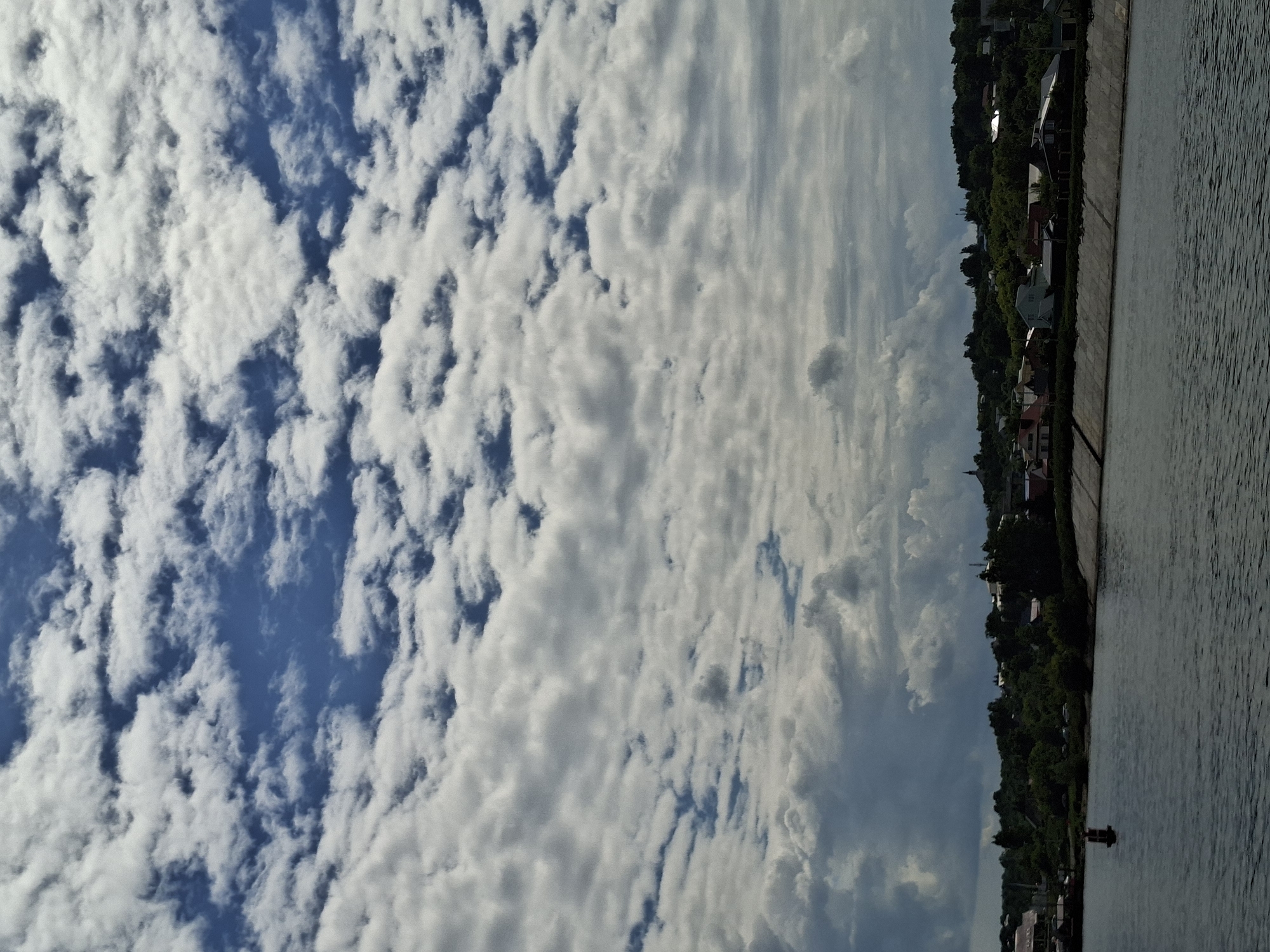 The image depicts a waterfront scene in Verhnij Uslon, Russia, characterized by a clear blue sky filled with fluffy white clouds on the left side and a more overcast, streaky cloud formation on the right. A body of water, likely a river, occupies the right side of the frame, its surface rippled by a gentle breeze. Along the embankment, a line of buildings and dense green trees suggests a residential or small town area. The architecture of the buildings appears varied, with some roofs visible and hints of various colors.
The perspective suggests the photo was taken from a boat or the water's edge, looking towards the shore. The lighting indicates it is daytime, with the sun likely high in the sky, casting soft shadows. The clouds suggest fair weather with potential for some cloud cover as the day progresses. The overall mood is peaceful and serene, with a sense of calm by the water. There is no visible text in the image. The scene captures a moment of natural beauty and tranquil village life in a Russian setting.