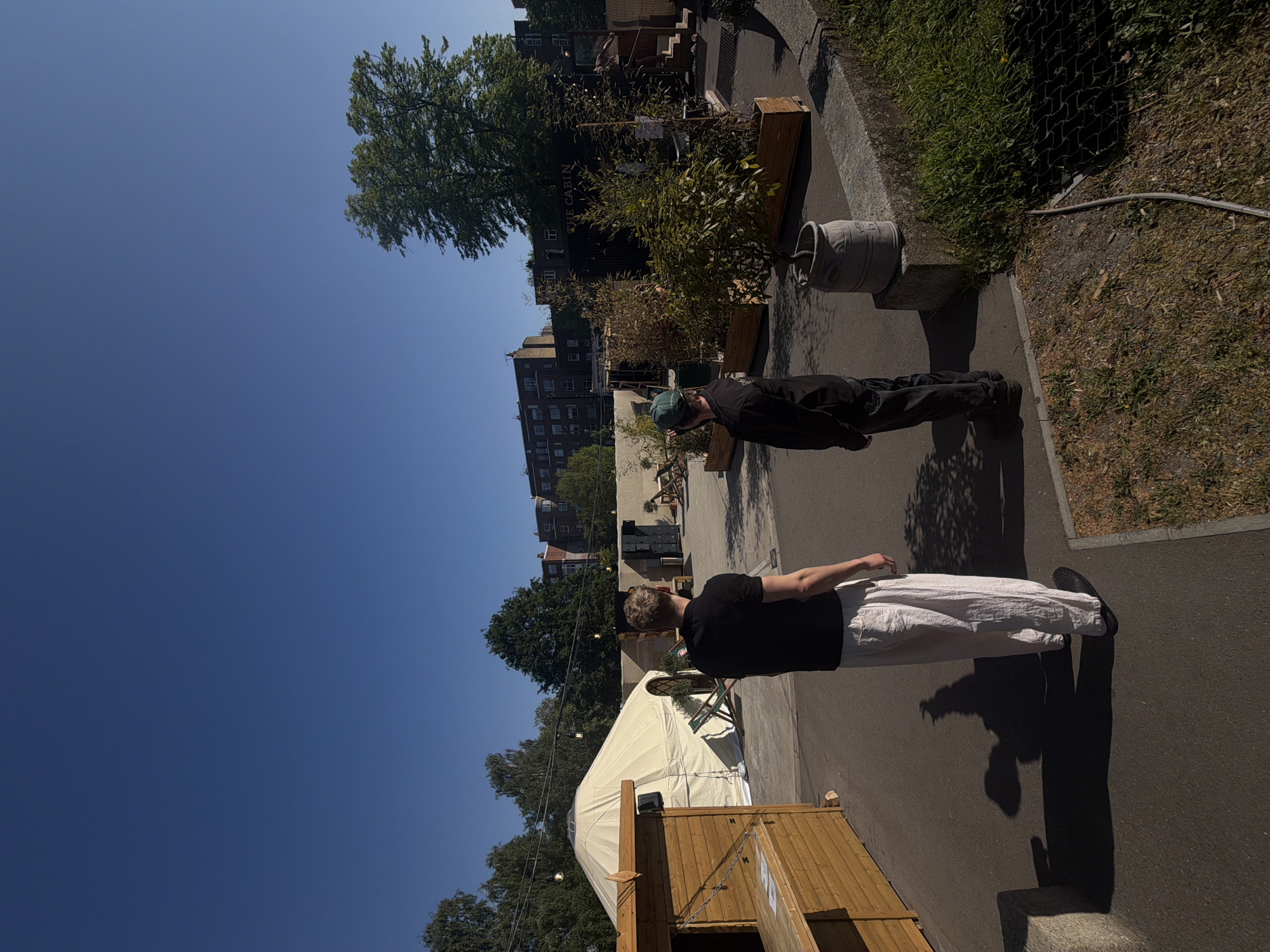 The image captures an outdoor urban scene in London, United Kingdom, bathed in bright, clear sunlight. Two individuals are visible on a paved surface, both casting long shadows indicative of late morning or afternoon. In the foreground, a person with light-colored hair, wearing a black t-shirt and white wide-leg trousers, stands with their back to the camera, looking towards the left. Further into the mid-ground, another person, dressed in a black long-sleeved top and a green baseball cap, stands facing away, looking towards the right.
To the left of the frame, a large white yurt or tent is set up, with a wooden structure, possibly a bar or booth, in the immediate foreground. In the mid-ground, behind the second person, a dark container-like building is visible, bearing the text "THE CABIN" on its side. Beyond this, several multi-story buildings typical of an urban setting can be seen under a vast, clear blue sky. The area is dotted with various potted plants and trees, and a grey bucket holding a plant sits near a metal fence and a patch of grass on the right. The overall setting suggests a pleasant outdoor gathering or event space.