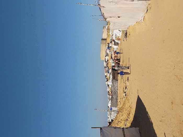 The image depicts a crowded, temporary settlement under a clear blue sky in Khan Yunis, Palestinian Territory. A wide, unpaved sandy path extends from the foreground into the mid-ground. On the left, a large mound of sand or dirt is visible, with a white tarp structure partially obscured by shadows in the lower left. On the right, several improvised shelters constructed from white tarps and supported by wooden poles line the path. In the mid-ground, a woman wearing a head covering and patterned clothing walks away from the viewer, accompanied by a young boy in a blue shirt and striped pants. Another young boy in a blue shirt walks further ahead on the right side of the path.
The background reveals a dense conglomeration of numerous makeshift tents and shelters, fabricated from various materials including tarps, fabric, and corrugated metal, spread across slightly elevated sandy terrain. Utility poles with wires are visible among these structures. Further back, on a raised mound, a prominent circular concrete or metal structure, possibly a water tower or reservoir, stands above the settlement. Distant figures and a red vehicle resembling a tractor are discernible within the camp area. The scene is illuminated by strong daylight, casting defined shadows.