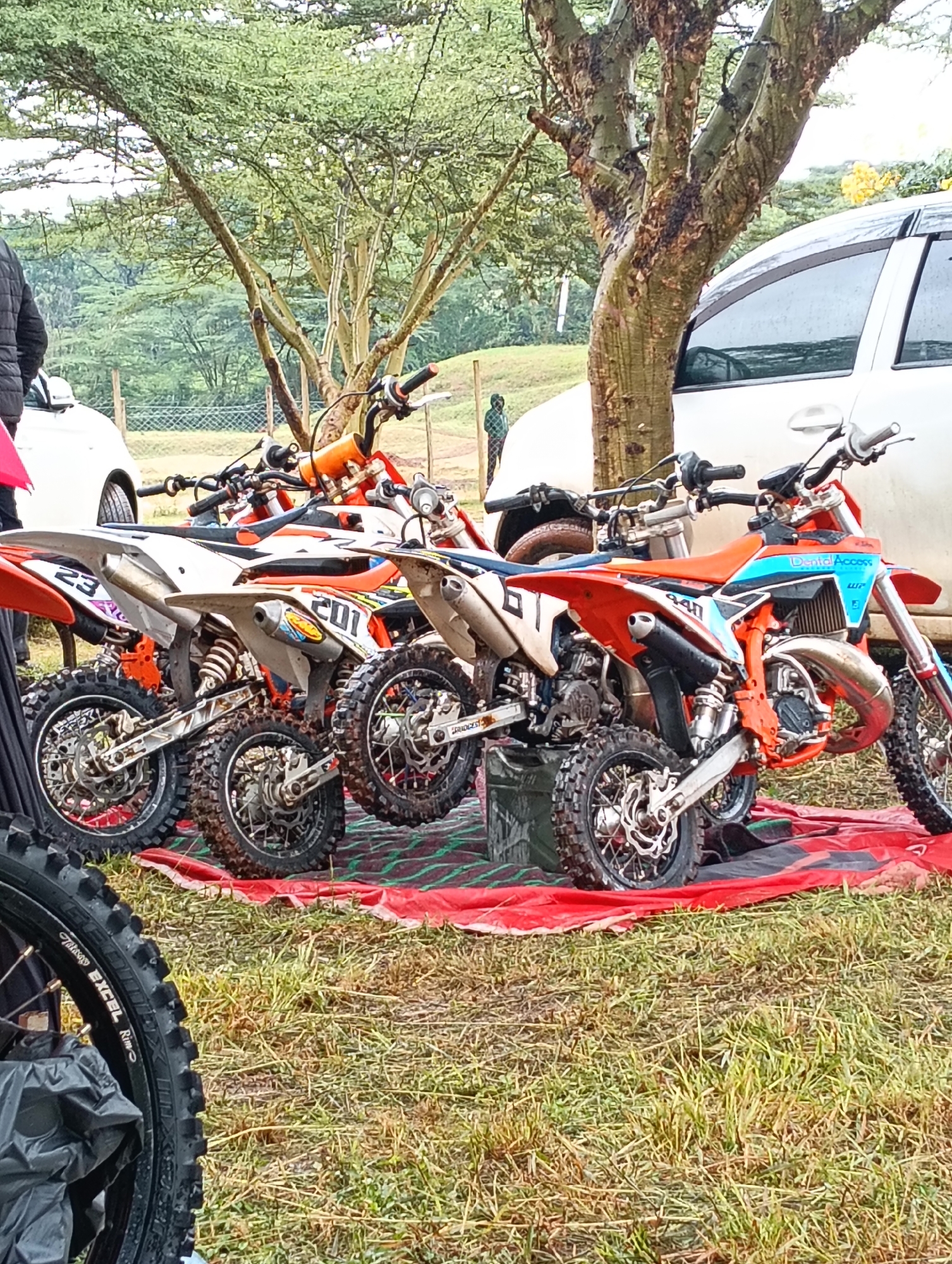 The image shows four small dirt bikes parked on a red tarp laid on a grassy field. The bikes are orange, white, and blue, and have racing numbers on them. The bikes appear to be KTM brand. A tree and a white car are visible in the background. A person wearing black jacket stands on the left, partially cut off from the image. The image appears to be taken outdoors during the day. The setting seems to be in a rural or semi-rural area, possibly near a park or open field. The exact location in Nairobi, Kenya cannot be determined with certainty from the image alone.