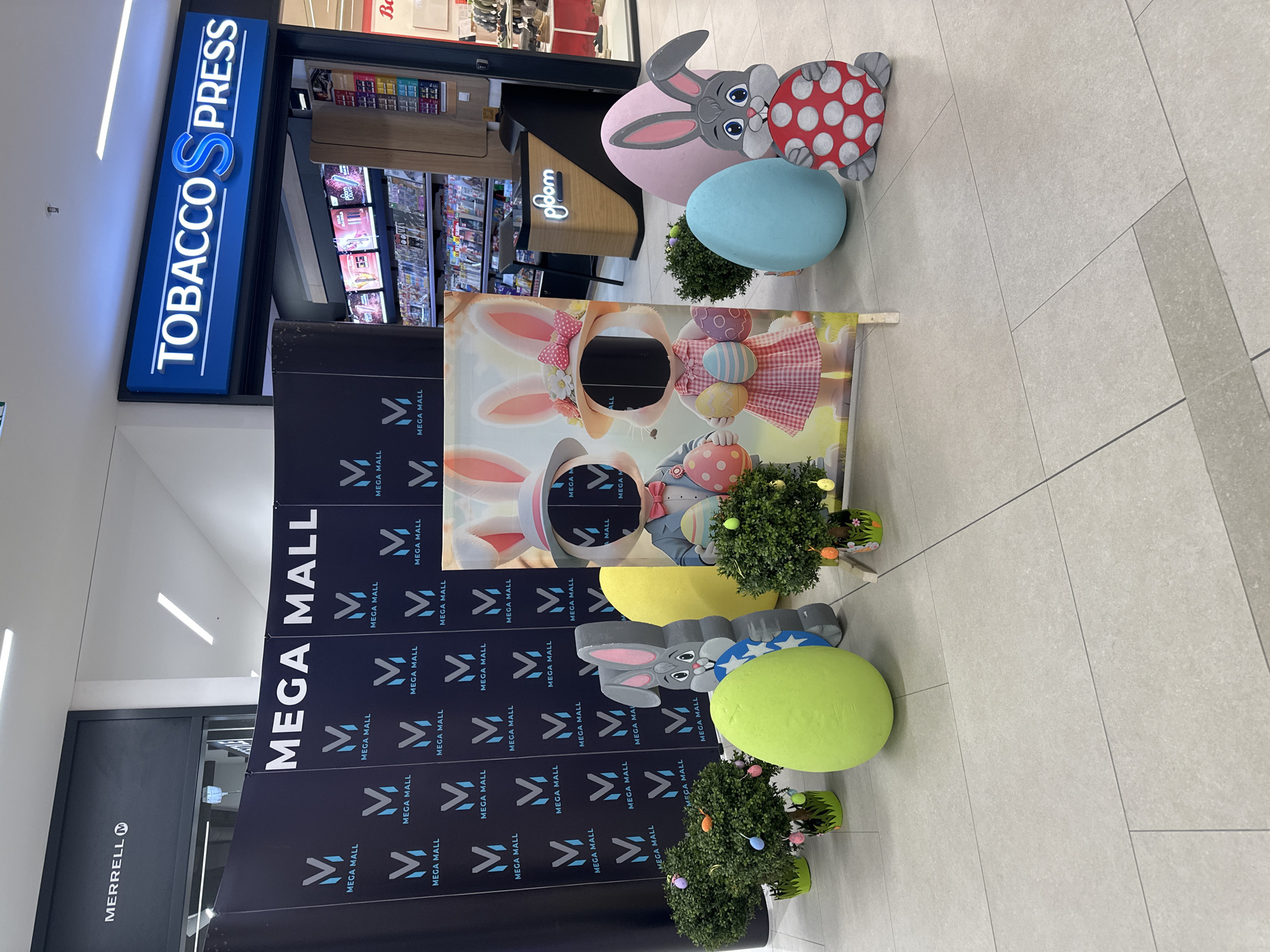 Here's a description of the image:
The image shows an Easter-themed photo opportunity set up inside the Mega Mall in Budva, Montenegro. In the background, storefronts can be seen, including a "Tobacco Press" shop and a "Merrell" store.
In the foreground, there is a backdrop with a cartoonish image of bunnies wearing hats and holding eggs, designed for people to pose behind with their faces in cut-out holes. This backdrop is decorated with "Mega Mall" logos. Beside the backdrop, there are decorative Easter displays, including oversized pastel-colored eggs and bunny figures, along with small potted plants. The floor is tiled.