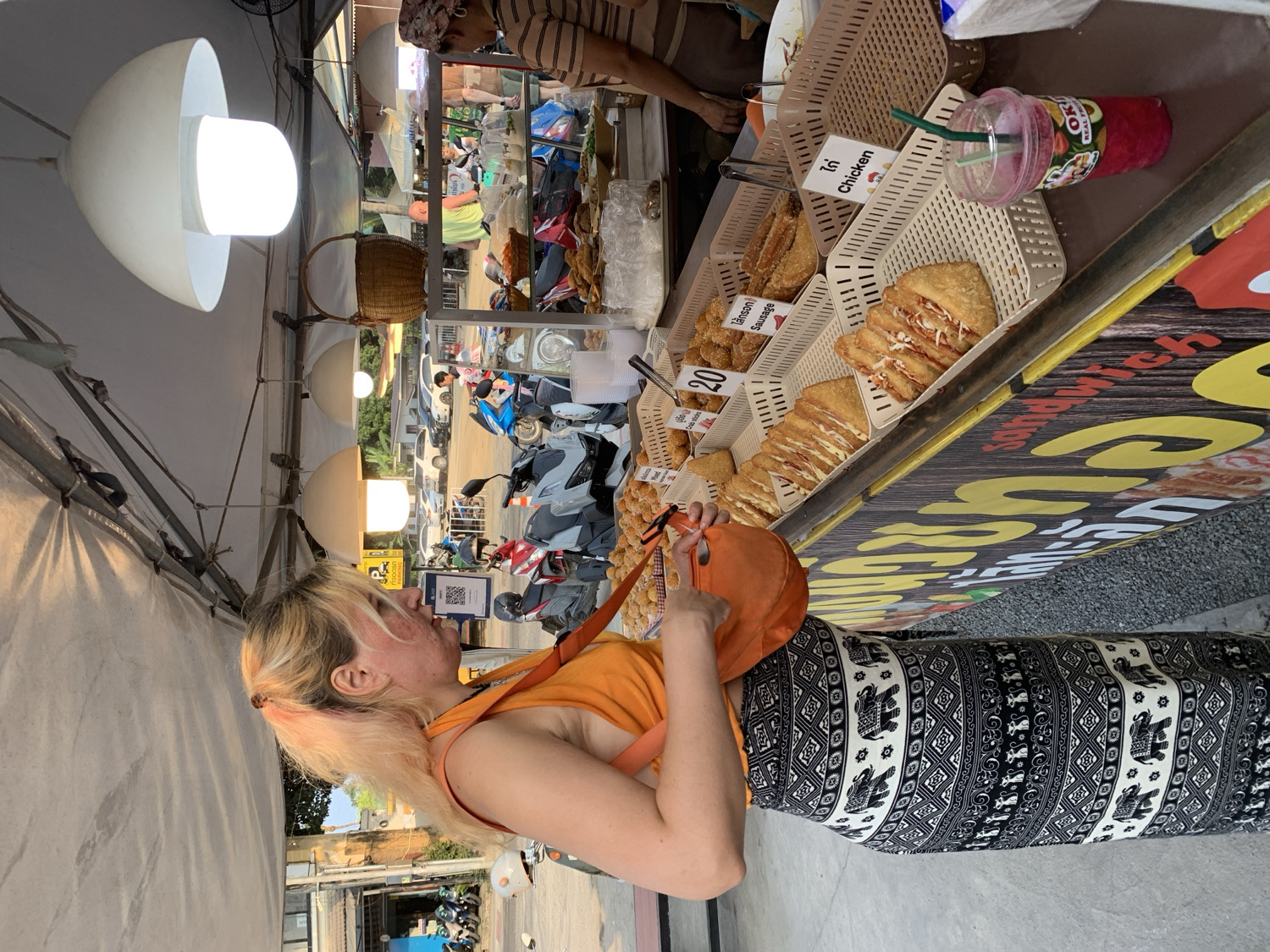 The image depicts an evening street food scene under a white canvas tent in บ้านบางรักษ์, Thailand. A blonde woman with pink highlights, wearing an orange tank top and a black and white patterned skirt/pants, stands in profile, attentively gazing at a food stall. She holds an orange shoulder bag and a smartphone displaying a QR code, likely for payment.
The food stall, illuminated by white mushroom-shaped lamps hanging overhead, showcases various baked and fried items arranged in plastic baskets. Visible labels indicate food types such as "ไก่ Chicken" and "ไส้กรอก Sausage," with one item priced at "20" (likely Thai Baht). A red or pink blended drink in a clear plastic cup with a green straw, labeled "OK! REAL FRUIT," rests on the counter. A vendor, partially visible behind the stall, attends to the offerings.
The stall's counter features a large banner with a wooden texture and prominent yellow lettering, including the partially visible word "Sandwich." In the background, parked motorcycles and scooters line a street, suggesting a busy local area. The artificial lighting confirms it is dusk or evening.