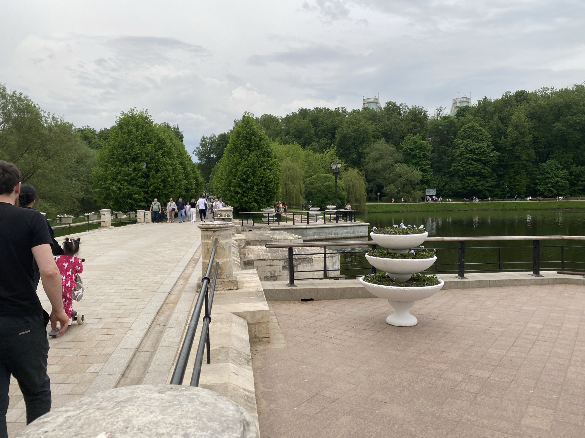 The image captures a scene in the park Tsaritsyno, Moscow, on a cloudy day. The foreground features a tiled walkway where a white, three-tiered planter with purple flowers sits. To the left, a pathway extends with a black railing and a low stone wall. Several people are walking along the path, including an adult and a small child wearing pink pajamas on a scooter. In the midground, a section of the pathway extends over a pond. On the other side of the pond, a line of trees stretches across the horizon. The towers of the Grand Palace can be seen among the trees.