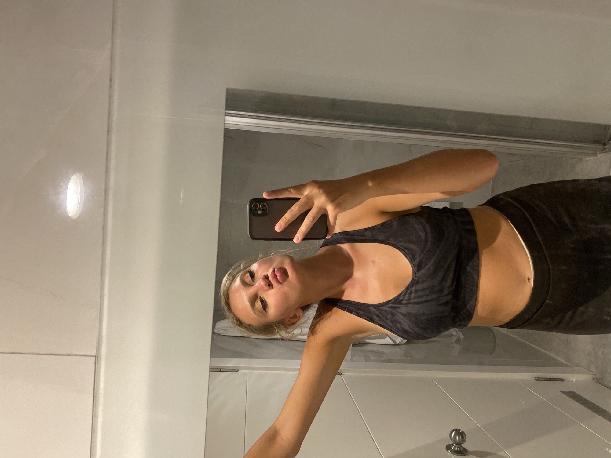 In a well-lit, modern bathroom with white tiled walls and a white ceiling, a young blonde woman is taking a selfie in a large mirror. She is wearing a black and gray patterned sports bra and black leggings. Her left arm is extended, holding her phone, and her lips are puckered in a kiss pose. Her right arm is bent at the elbow, with her hand resting on her hip. The bathroom appears to have a glass shower door to her right and a white cabinet door to her left, with a silver handle. The floor is a light gray tile. The overall impression is a casual, personal moment captured in a private space, likely a hotel bathroom in Aksu, Turkey, given the context of the prompt.