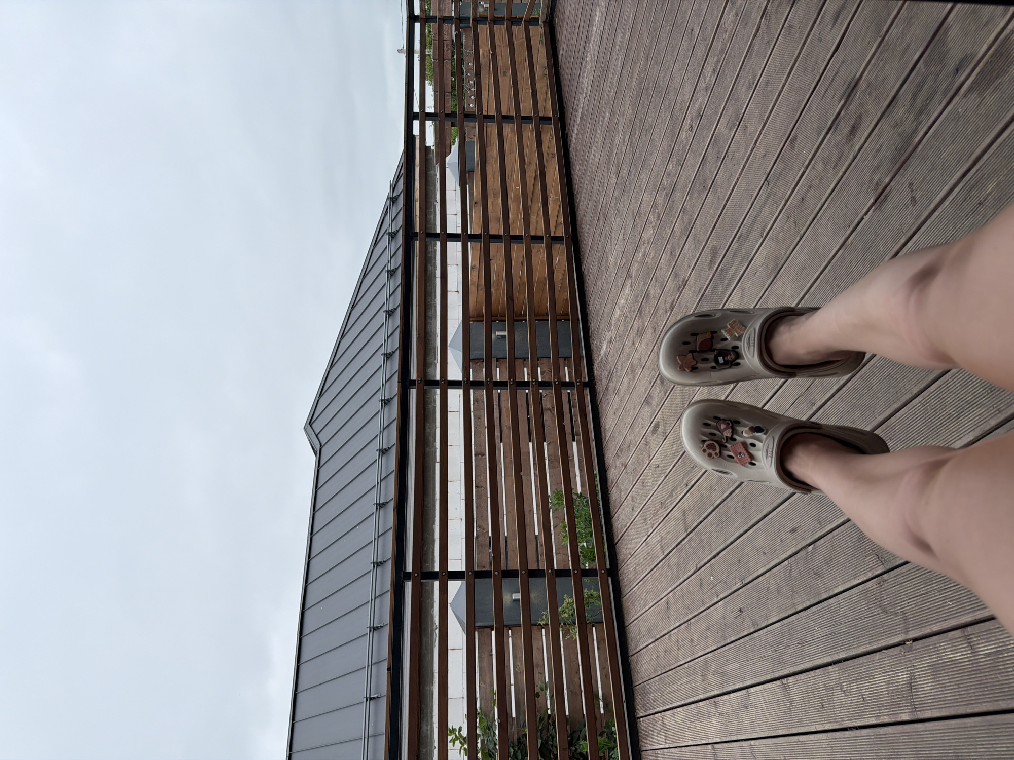 A person's legs, wearing beige crocs with charms, are stretched out on a wooden deck. The deck is made of parallel wooden planks. In the background, a modern house with a dark metal roof is visible. A wooden fence with horizontal slats runs along the edge of the deck. The sky is overcast. The image appears to have been taken from a seated or reclining position. The location is identified as Semiluki, Russia.