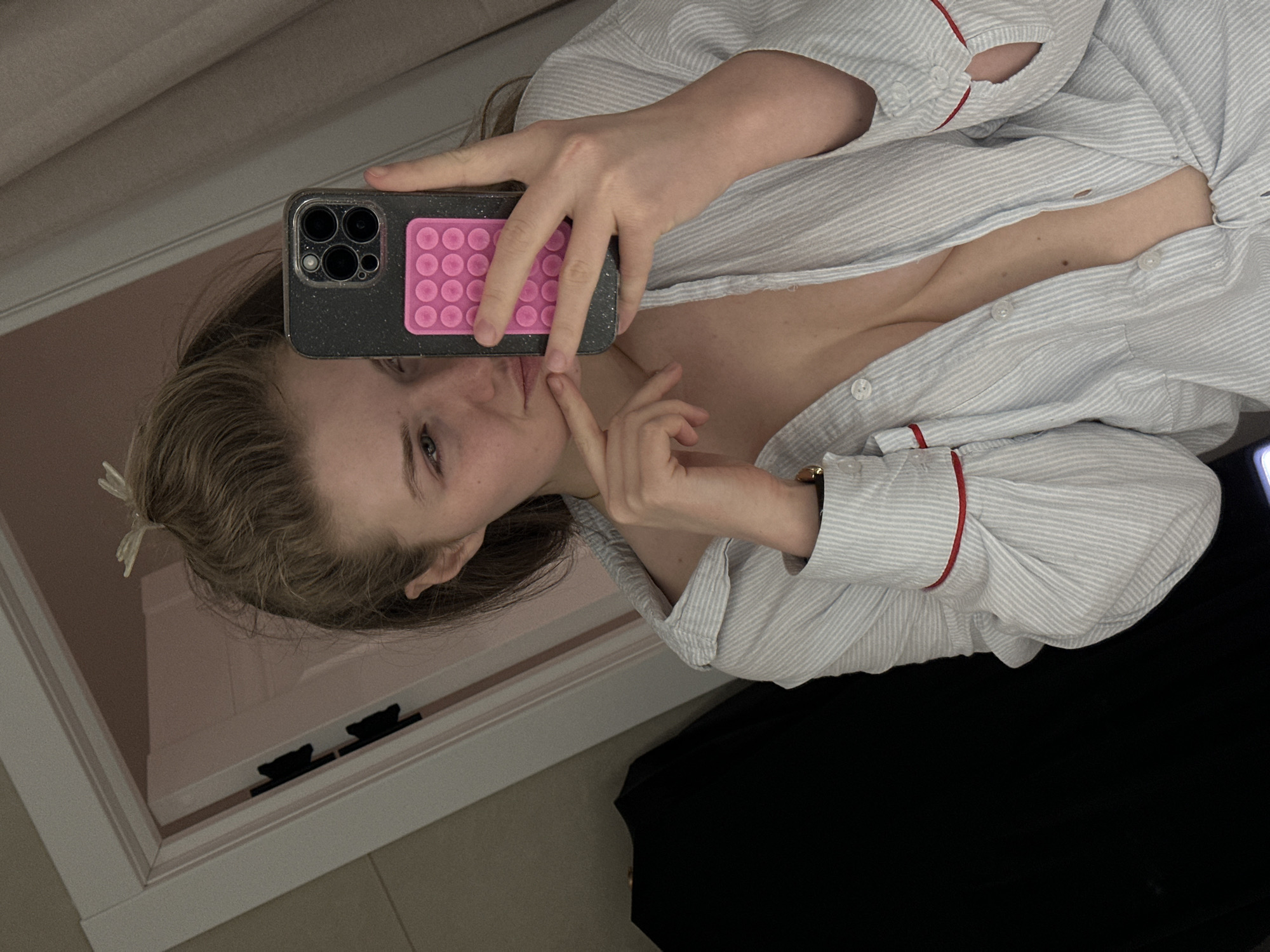 A young woman is taking a selfie in what appears to be a closet. She is holding a smartphone with a speckled case and a pink pop-socket on the back. The phone obscures part of her face. She is wearing a striped shirt with red trim. The background shows a closet with a white door and black bear-shaped pulls. There is no indication of the city of Kazan, Russia in this image.