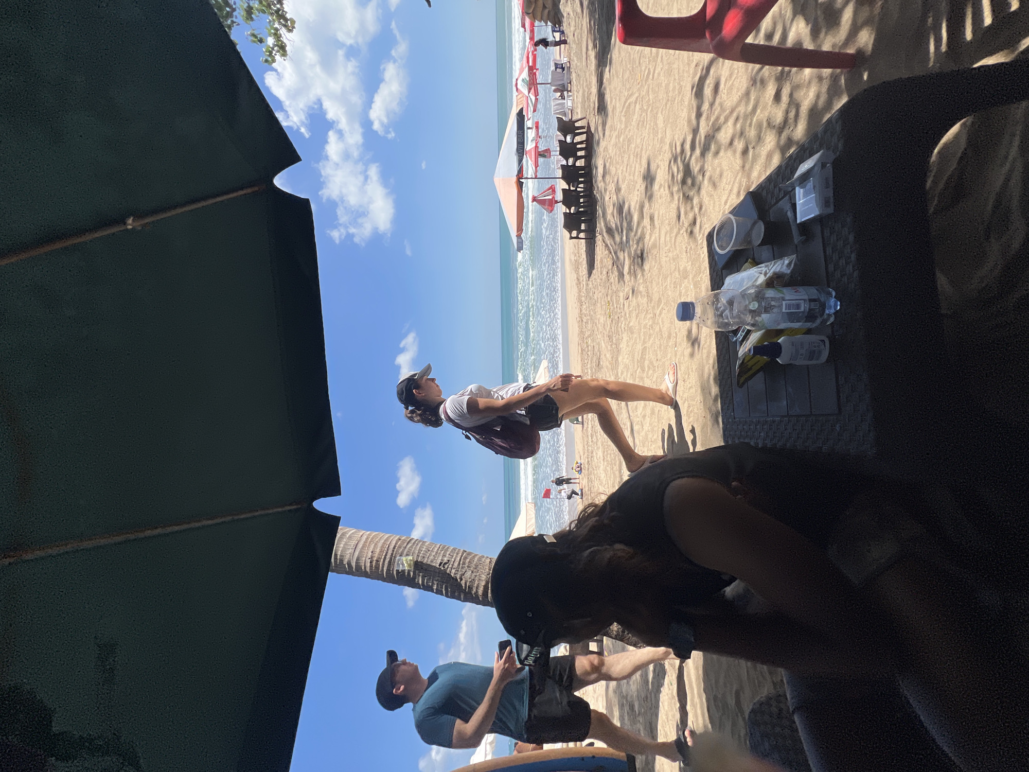 In Kuta, Indonesia, on a sandy beach under a green umbrella, two men and two women are visible. The man on the left, wearing a blue t-shirt and black shorts, walks to the left, holding a smartphone. A woman in a white t-shirt and black shorts walks to the right, with a maroon backpack. In the foreground, a person wearing a black t-shirt and a black cap is seated facing away from the camera, looking down at a black table. On the table are a plastic bottle of water, a cup, a cigarette pack, and other small items. In the background, the ocean waves crash onto the shore, and beach umbrellas are set up. The sky is blue with scattered clouds.