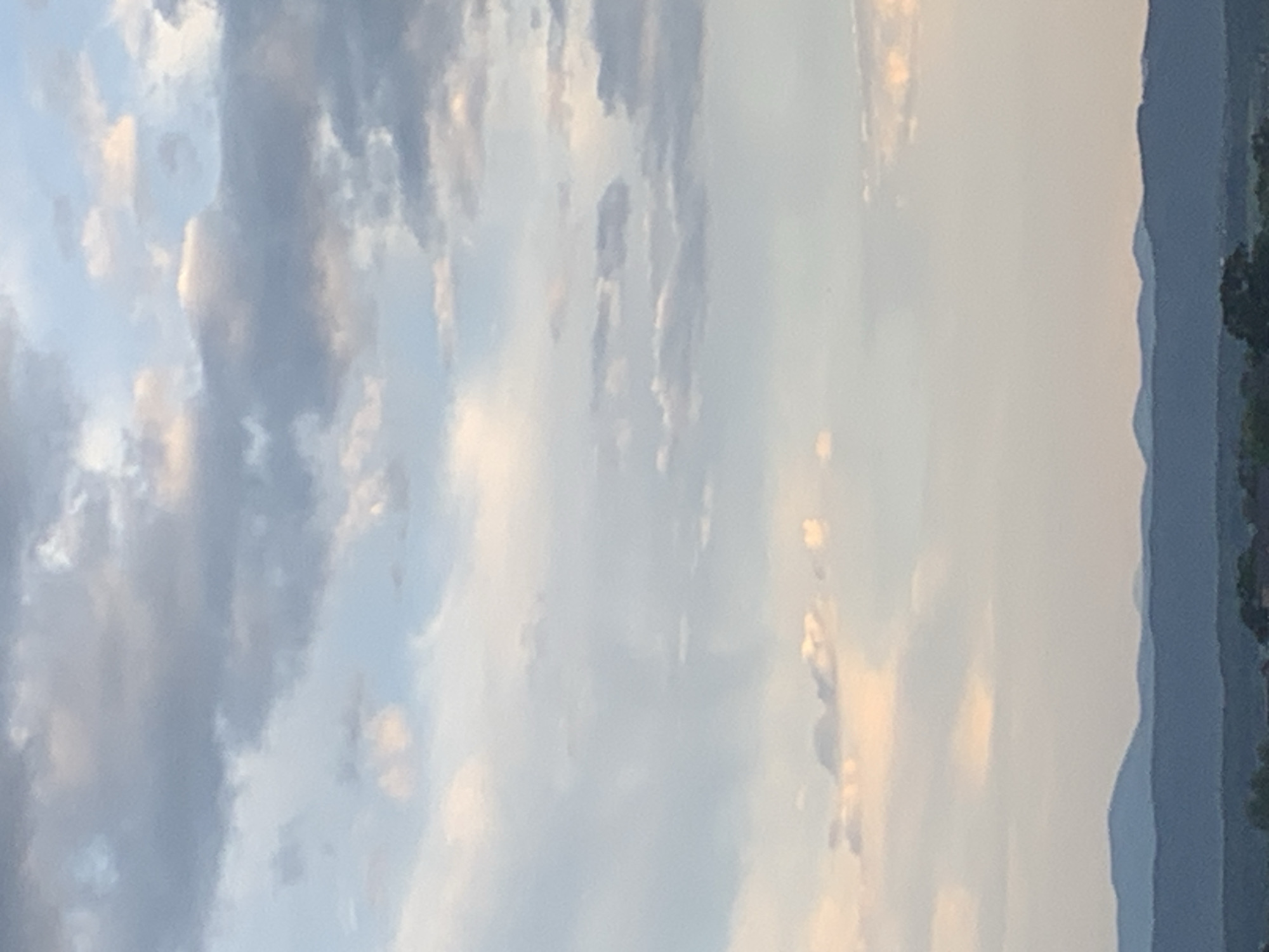 This is a high-angle, vertical shot of a sky with scattered clouds over a distant, dark, tree-lined mountain range. The sky is a pale blue with wispy, white and grey clouds, some tinged with pink from the setting sun. The mountain range is dark blue and appears to be composed of several layers of hills. The bottom of the image shows a sliver of a dark green, possibly forested, landscape with some buildings visible. There are no people or specific identifiable landmarks of Kolomyia, Ukraine in this image. The image captures a natural landscape scene.