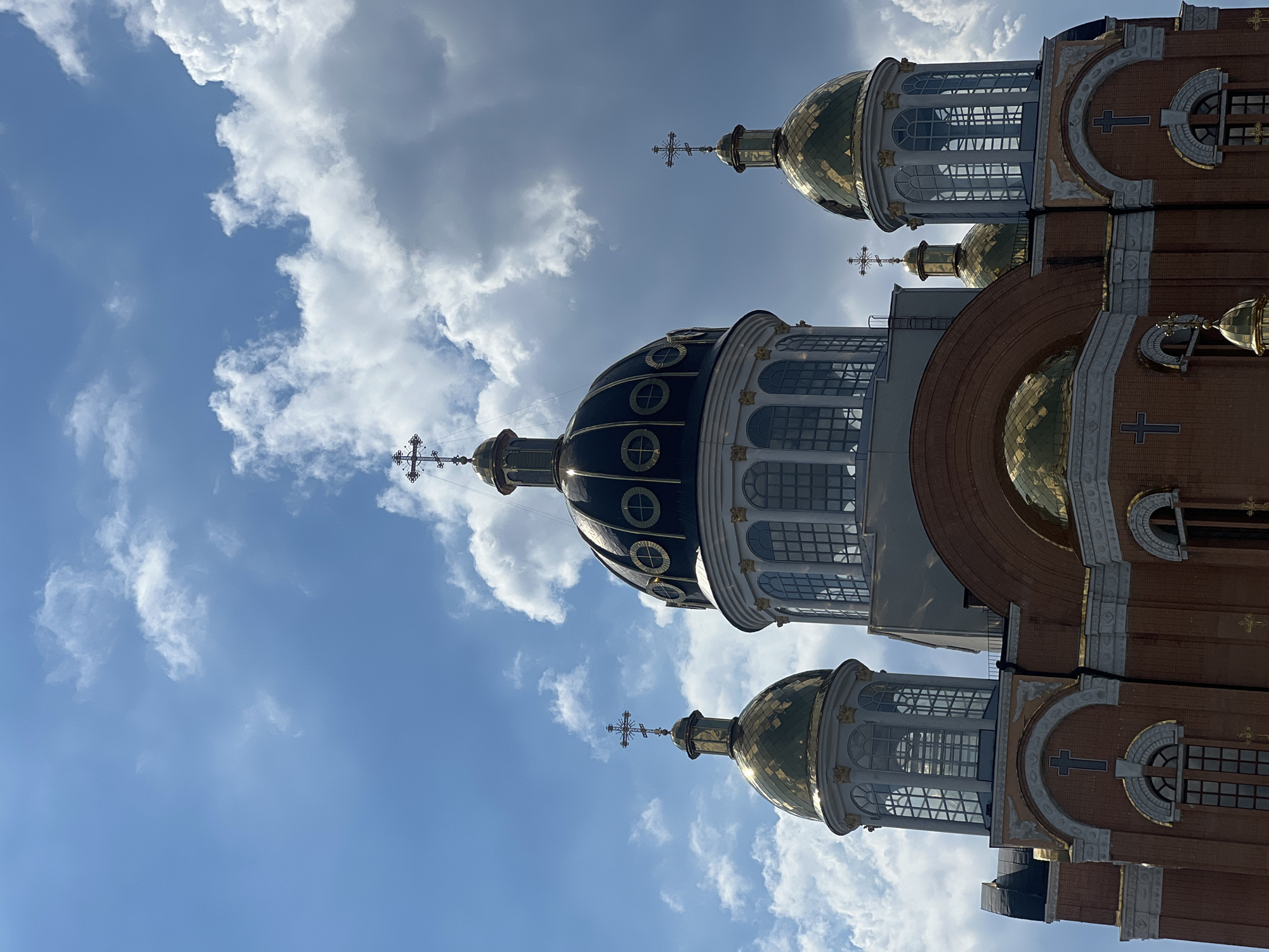 The image shows a low-angle shot of the exterior of the Ascension Cathedral, a large Eastern Orthodox church located in Kyiv, Ukraine. The church features multiple golden domes, with the central dome being the largest and adorned with a cross. Several smaller golden domes are visible surrounding it, also topped with crosses. The main structure of the building is constructed with red brick, with white architectural details and ornate windows. The sky above is a vibrant blue, dotted with fluffy white clouds, suggesting a sunny day. The image is a close-up, focusing on the upper portions of the cathedral, highlighting its impressive architecture and religious symbolism against the backdrop of the sky. No people are visible in the frame.