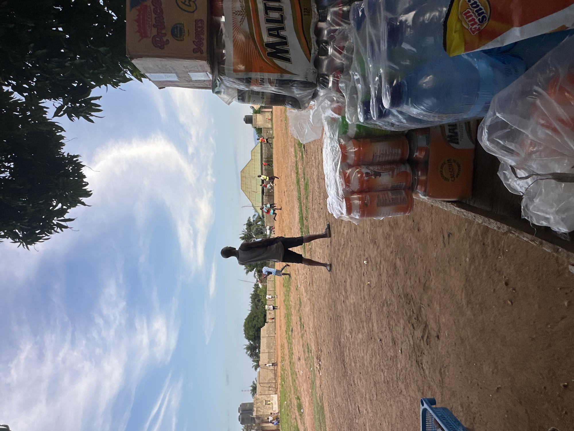 This image depicts an outdoor scene, likely in Akwanga, Nigeria, with a focus on a makeshift stall selling various beverages and snacks. The stall is filled with bottled water, canned drinks, and packaged snacks. Prominent text on some items includes "MALT," "Princess," and "Fun Snack."
In the background, several people are visible in a dusty, open field. Some appear to be engaged in informal activity, possibly a game, given their scattered positions and dynamic poses. The terrain is dry and uneven, suggesting a rural or semi-urban setting. Beyond the field, there are low-rise buildings and trees, with a clear sky above.
The lighting suggests it is daytime, likely late afternoon, with the sun casting some shadows. The sky is partly cloudy with wispy cirrus clouds. The overall atmosphere seems casual and active, with the presence of people enjoying an outdoor space. The foreground is dominated by the stall's merchandise, creating a sense of commerce and everyday life.