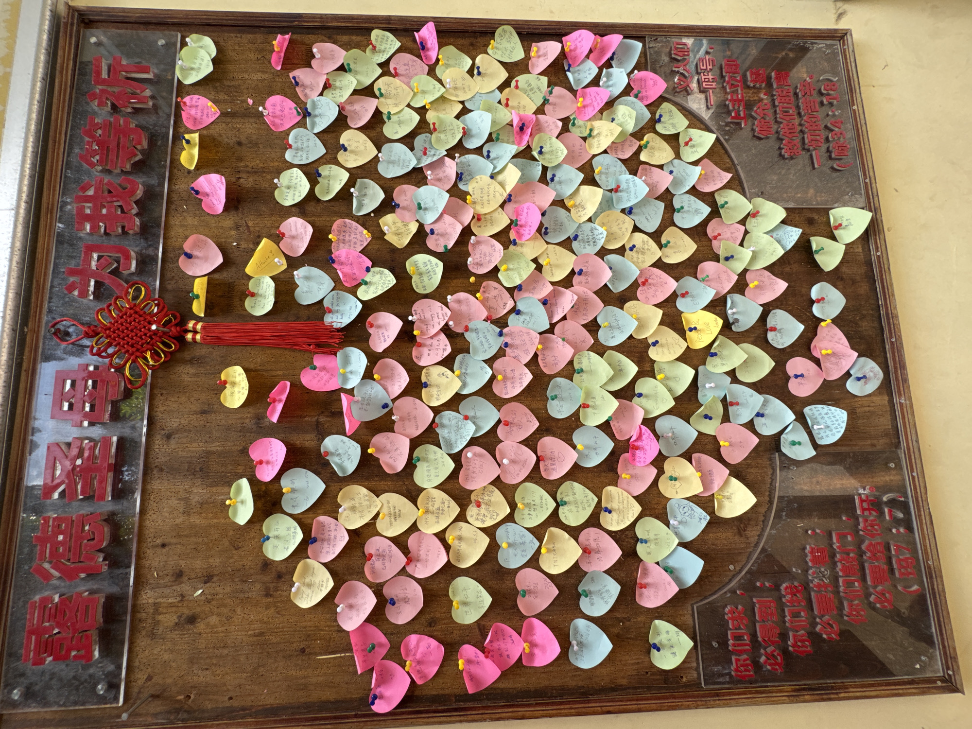 The image captures a framed wooden board densely covered with colorful, heart-shaped paper notes, each attached with a pushpin. The notes are in various shades of pink, yellow, green, and blue. There appear to be writings in Chinese on the notes. A red Chinese knot is affixed to the left side of the board. Two transparent plaques with red Chinese characters are mounted on the left and right sides of the board. The lighting appears to be natural, creating subtle shadows across the surface. The board is located in the Liwan District, China.