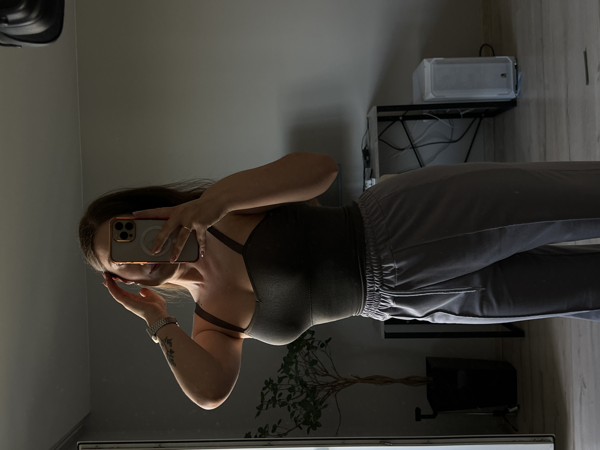 A young woman with long brown hair stands in front of a mirror, taking a selfie. She is wearing a dark olive green tank top and light grey sweatpants. Her left arm is raised, and her hand is touching her head, obscuring her face. A silver watch is visible on her left wrist. The background shows a grey wall, a potted plant to the left, and a desk with a computer tower and cables to the right. The floor appears to be wood. The lighting suggests it is daytime, with a strong light source casting shadows. The image is set in an indoor residential space, likely an apartment or house.