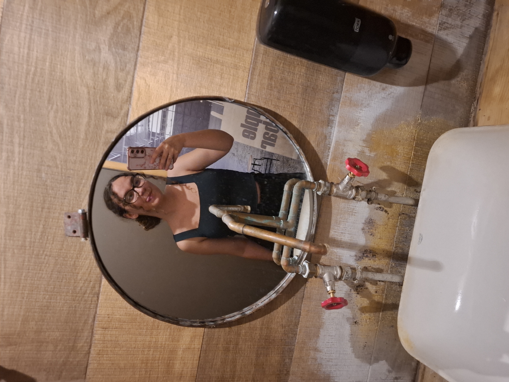 A young woman with curly brown hair and glasses is reflected in a round mirror, taking a selfie with her phone. She is wearing a dark tank top and patterned pants. She is sticking her tongue out. The mirror is framed by a weathered metal circle and attached to a wooden wall with a rusty hinge. Below the mirror, a white sink is visible with exposed copper pipes and red valve handles. A black soap dispenser is mounted on the wall to the right of the sink. The background in the mirror shows part of a building interior with a sign that is partially visible. The overall setting appears to be a rustic restroom.